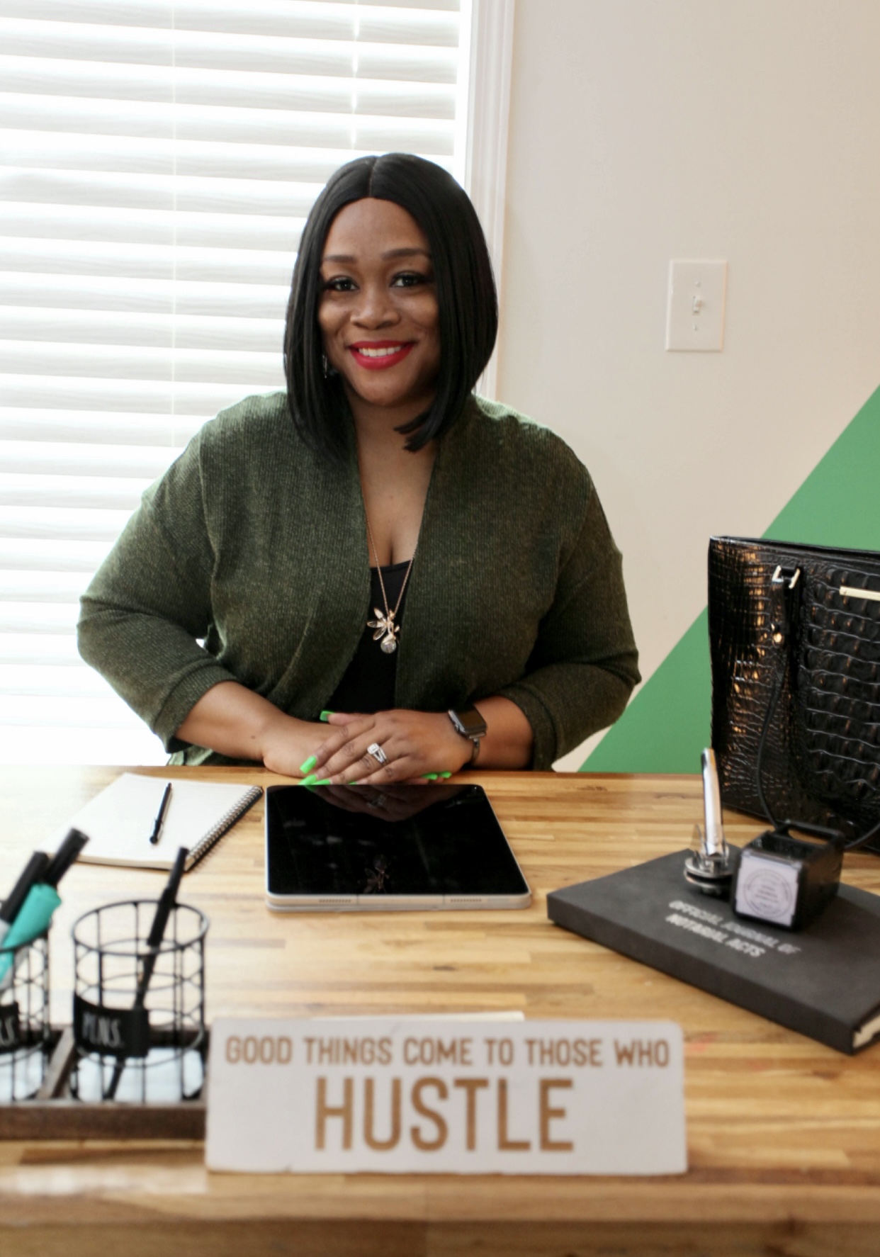 Woman with shoulder-length black hair smiling, sitting at a desk with a sign that says 'HUSTLE'.