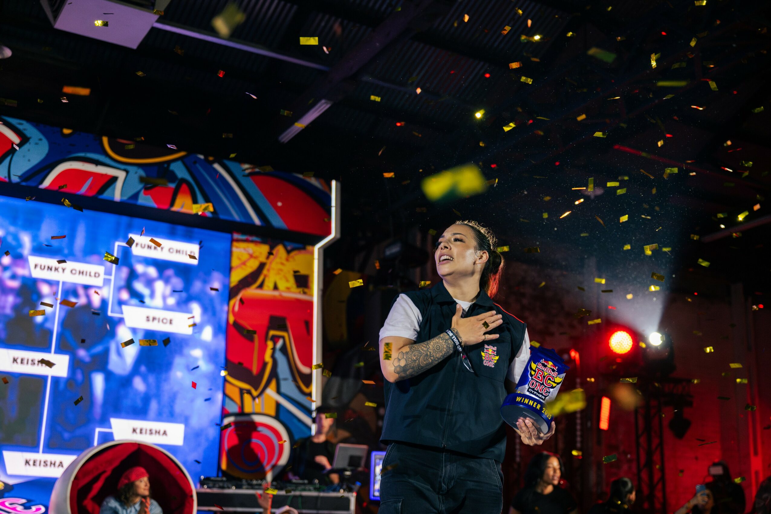 Woman on stage holding a trophy, with a colorful screen and confetti falling around her.