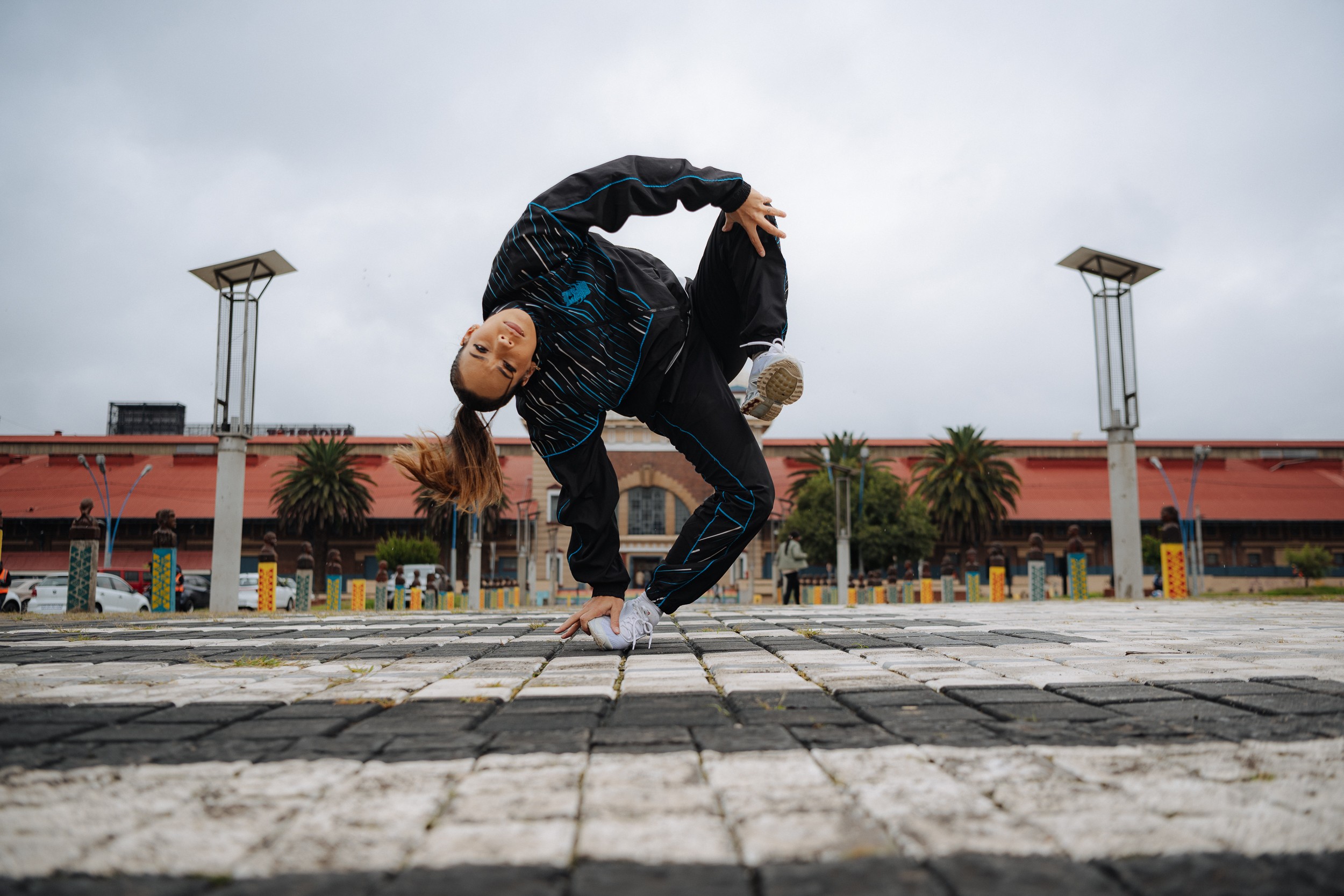 Person performing a breakdance move outdoors on a paved area with a building and palm trees in the background.