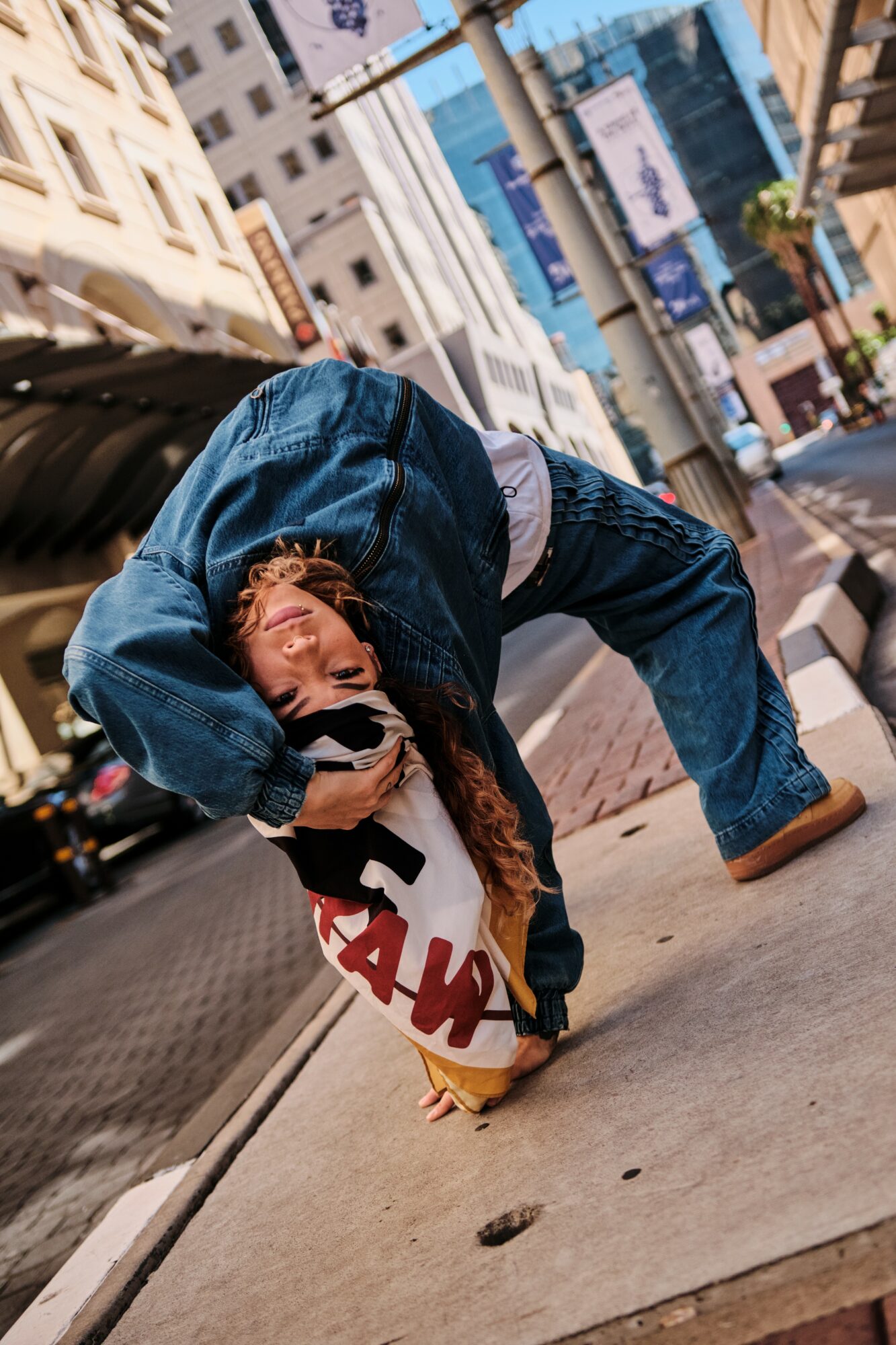 Person bending over on city sidewalk holding a bag, buildings and street scene in background.