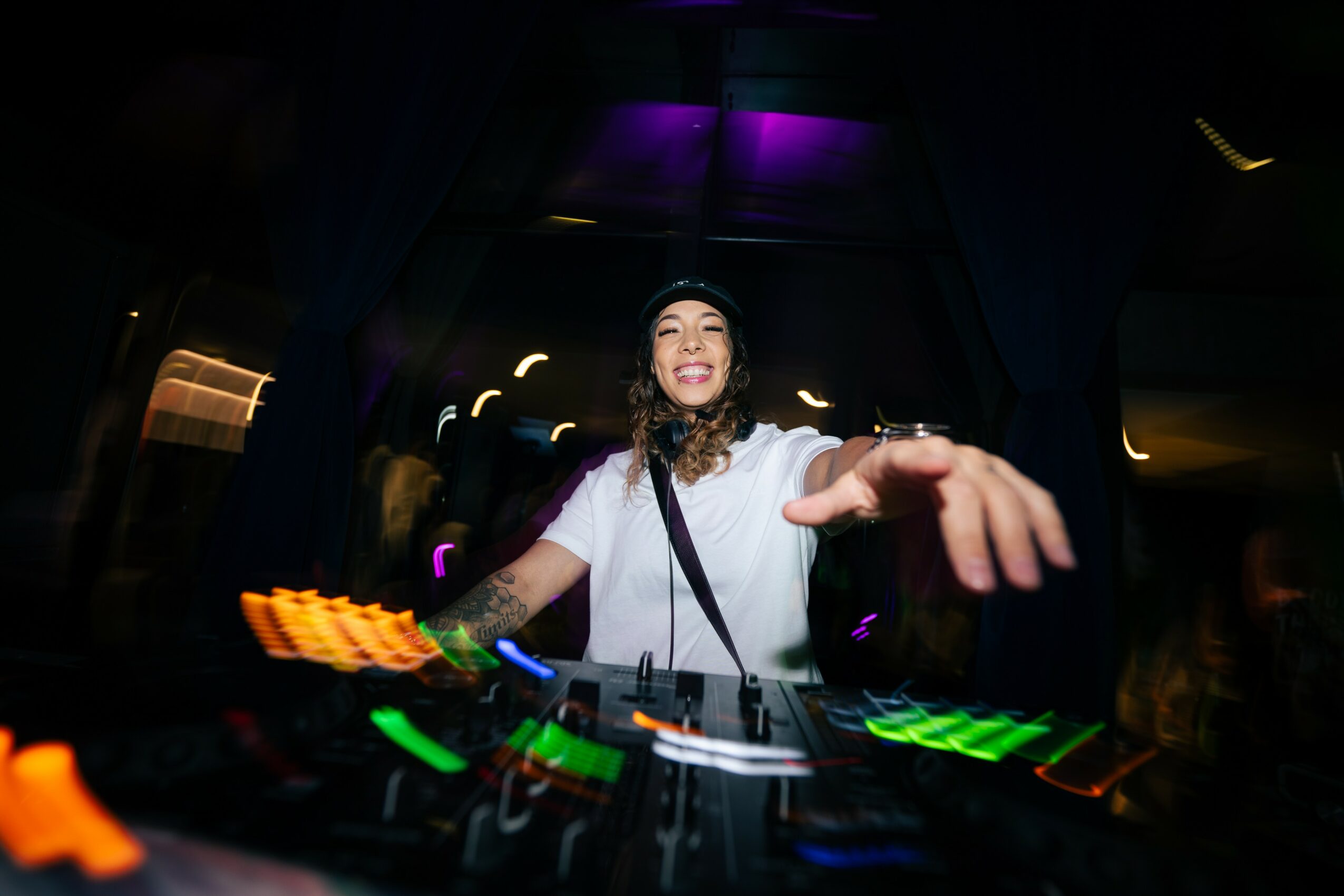 Female DJ smiling and reaching out at a DJ booth with colorful equipment in a dark club setting.