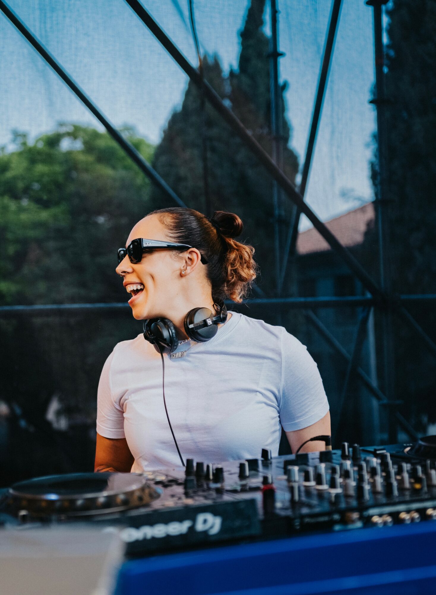 Female DJ wearing sunglasses and headphones, smiling, in front of DJ equipment outdoors.