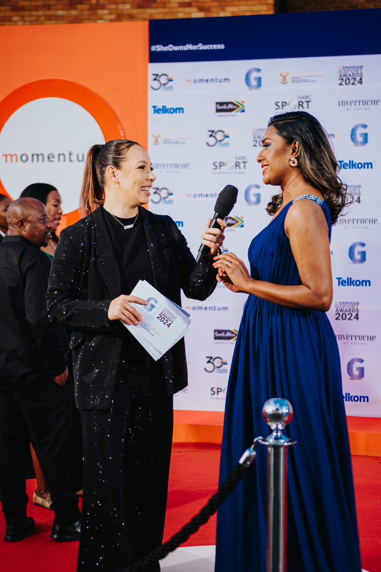 Two women on stage, one holding a microphone and paper, the other in a blue dress, with a backdrop of logos.