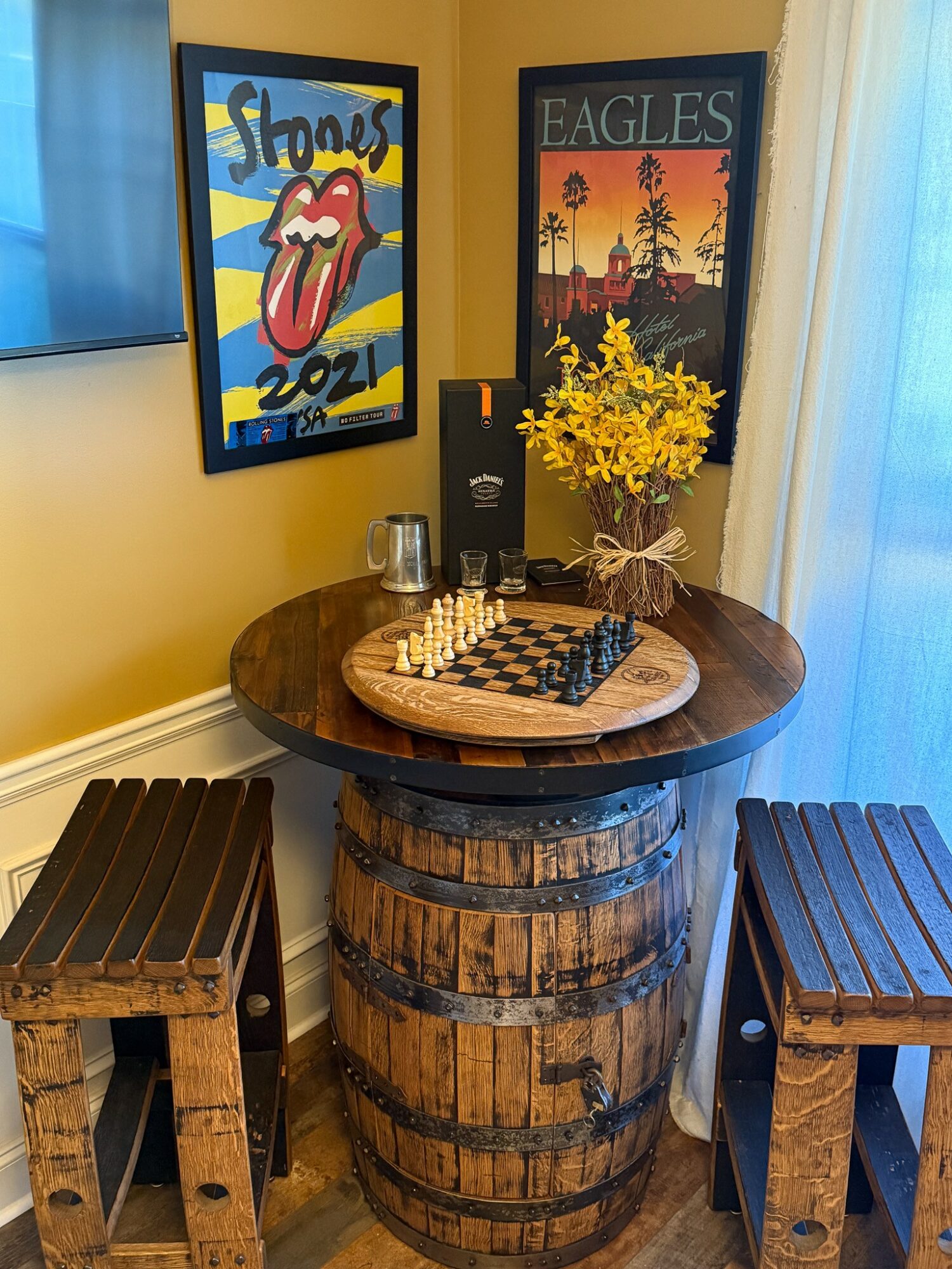Round wooden table with chessboard, yellow flowers in a vase, and two glasses, flanked by two wooden stools, in a room with posters on the wall.