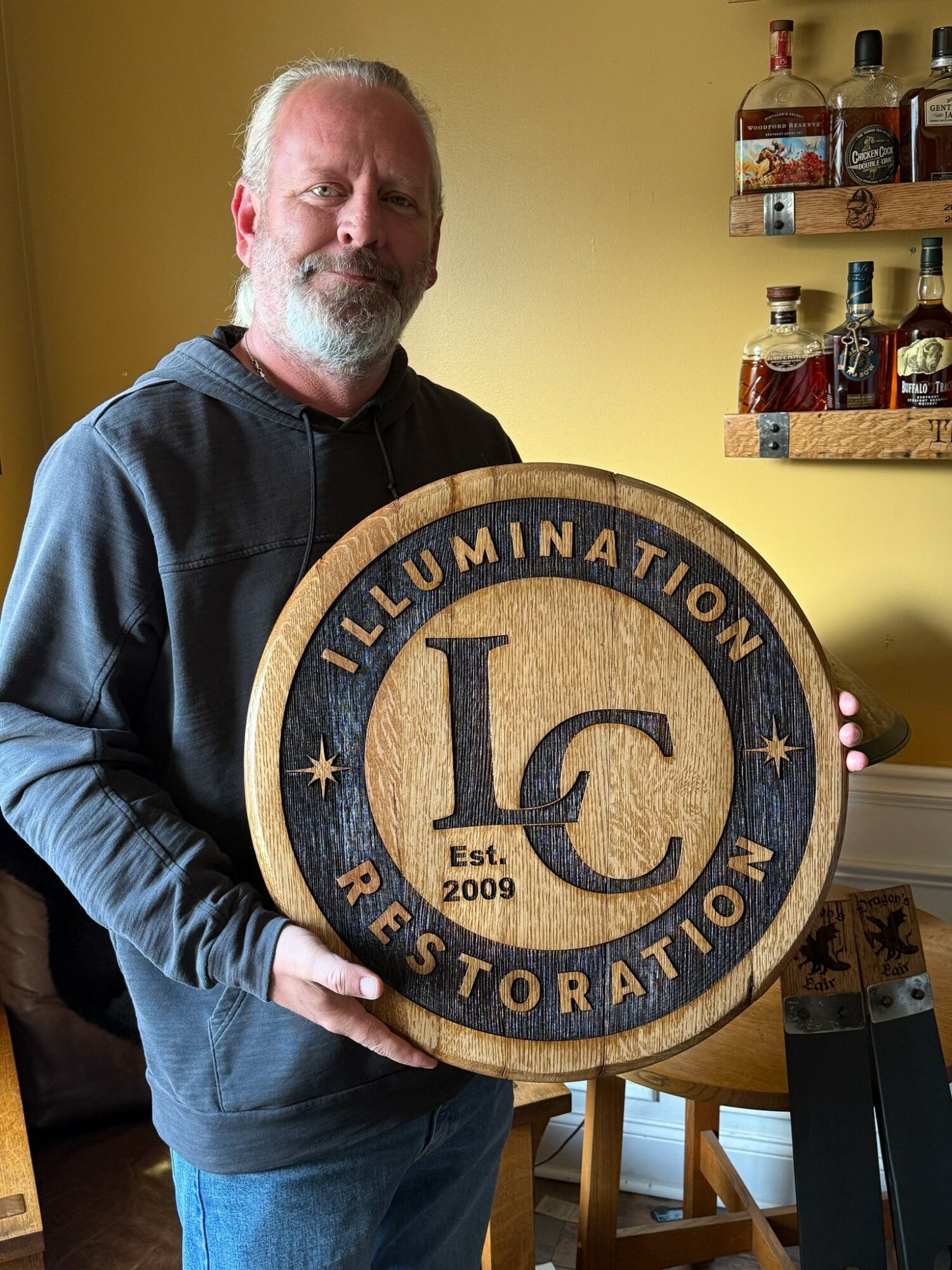 Man with gray beard holding a circular sign with 'LC' and 'Est. 2009' in a room with shelves of bottles.