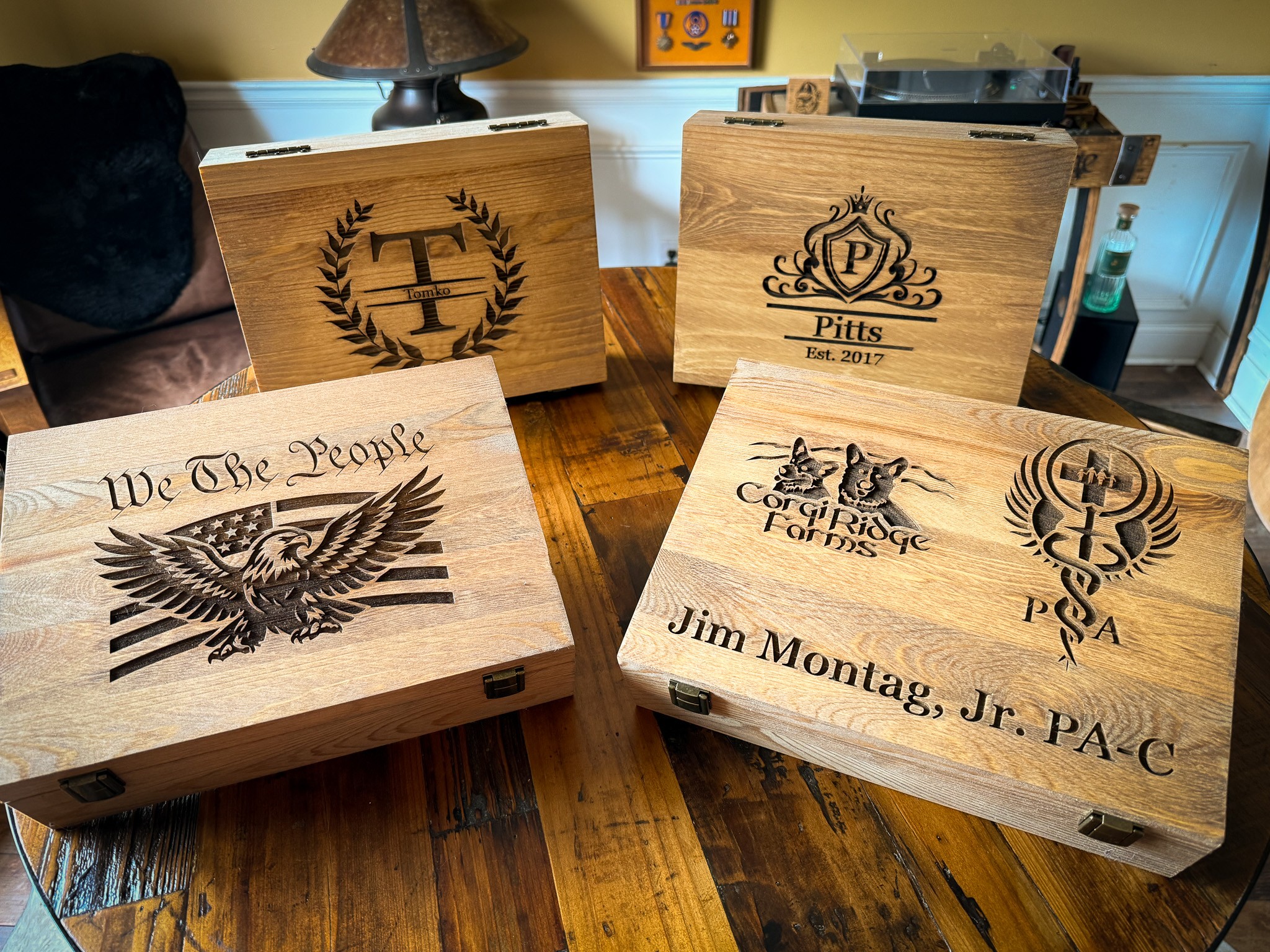 Wooden boxes with engraved logos and text, placed on a wooden table in a room with a lamp and wall decor.