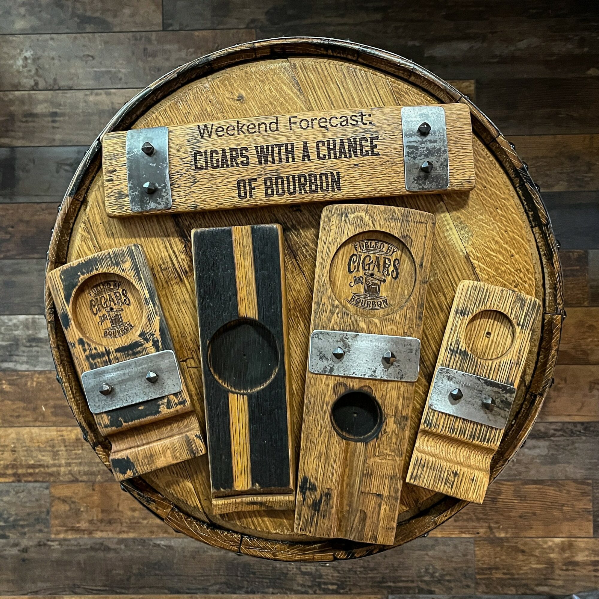 Wooden barrel top with four wooden and metal signs promoting bourbon and cigars, and a black and gold striped card holder.