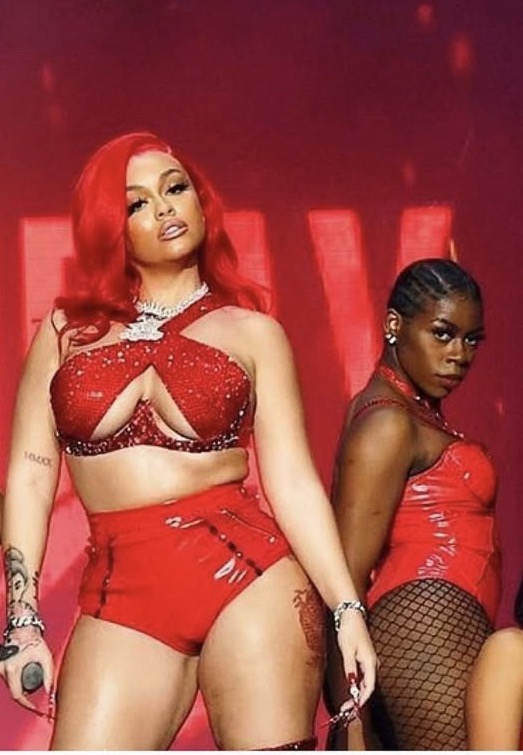 Two women in red outfits perform on stage with red lighting in the background.