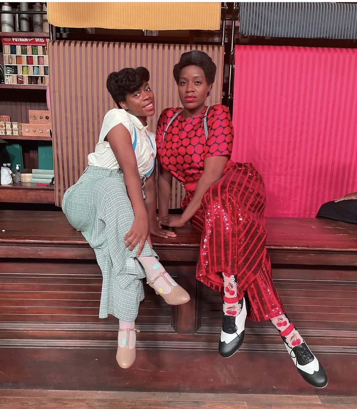 Two women sitting on a wooden bench indoors, with colorful background elements and shelves behind them.