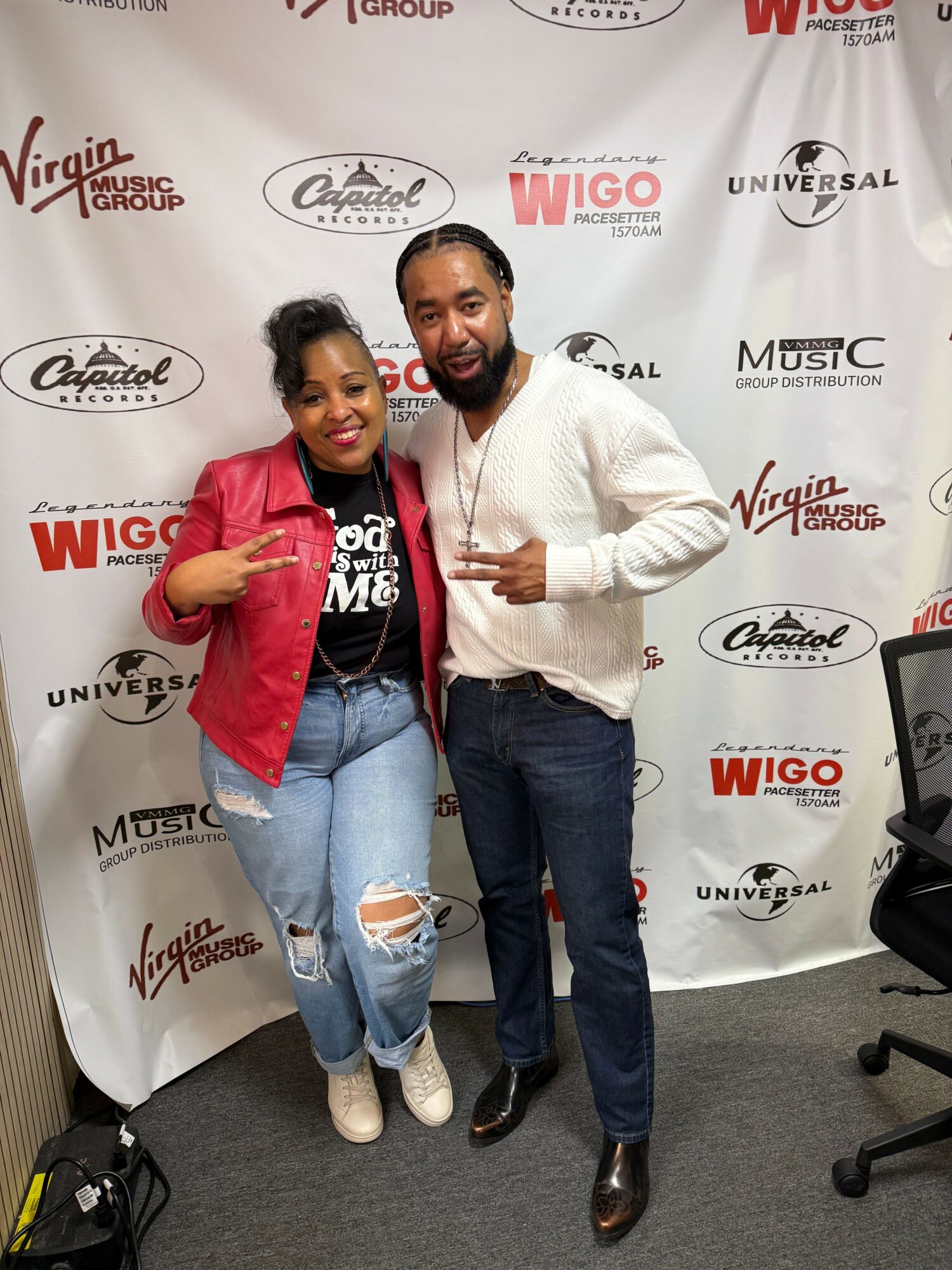 Two people posing in front of a backdrop with logos, smiling, making peace signs, one wearing a red jacket and ripped jeans.
