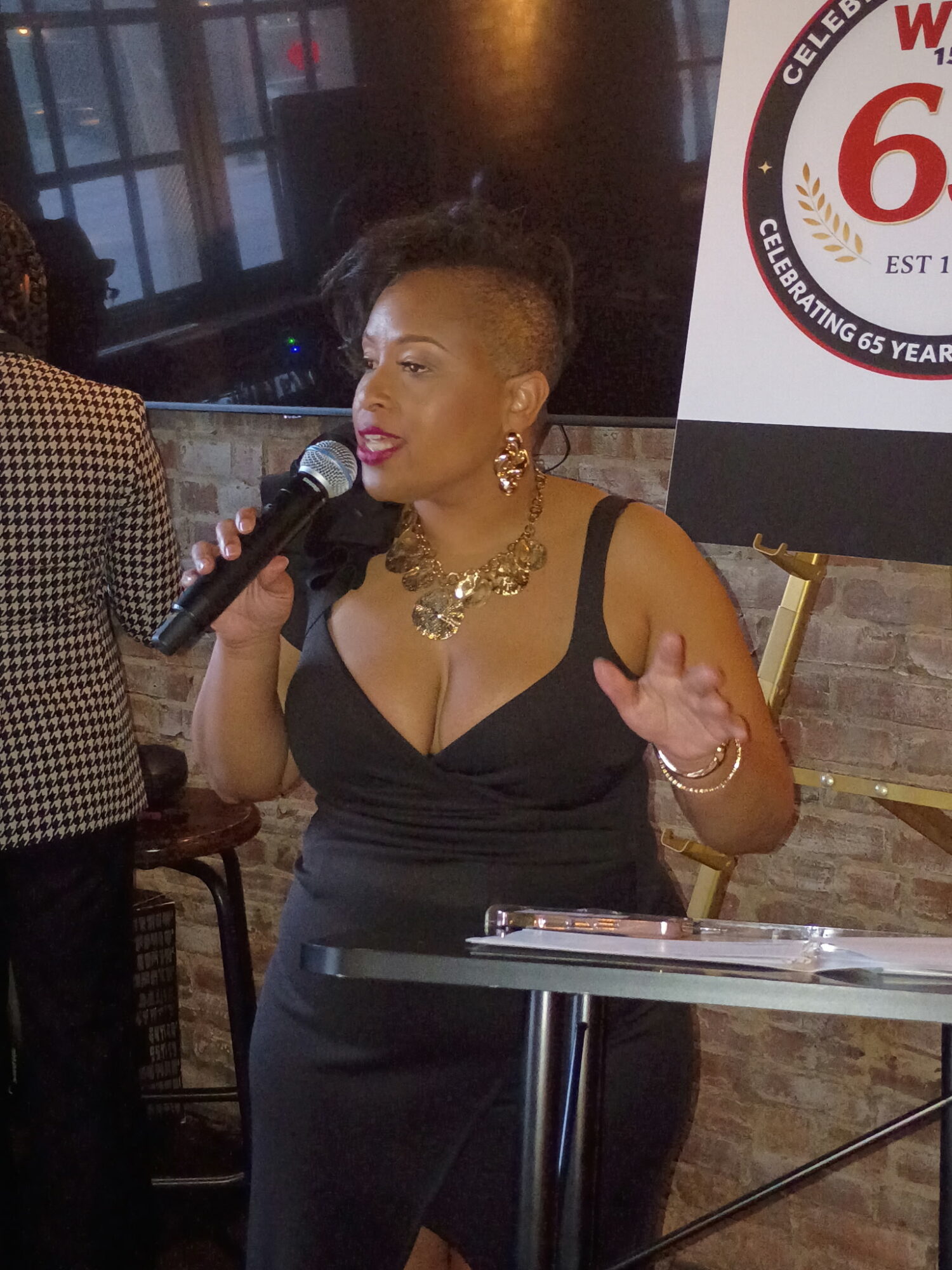Woman speaking into microphone, wearing black dress and jewelry, seated at a table indoors.