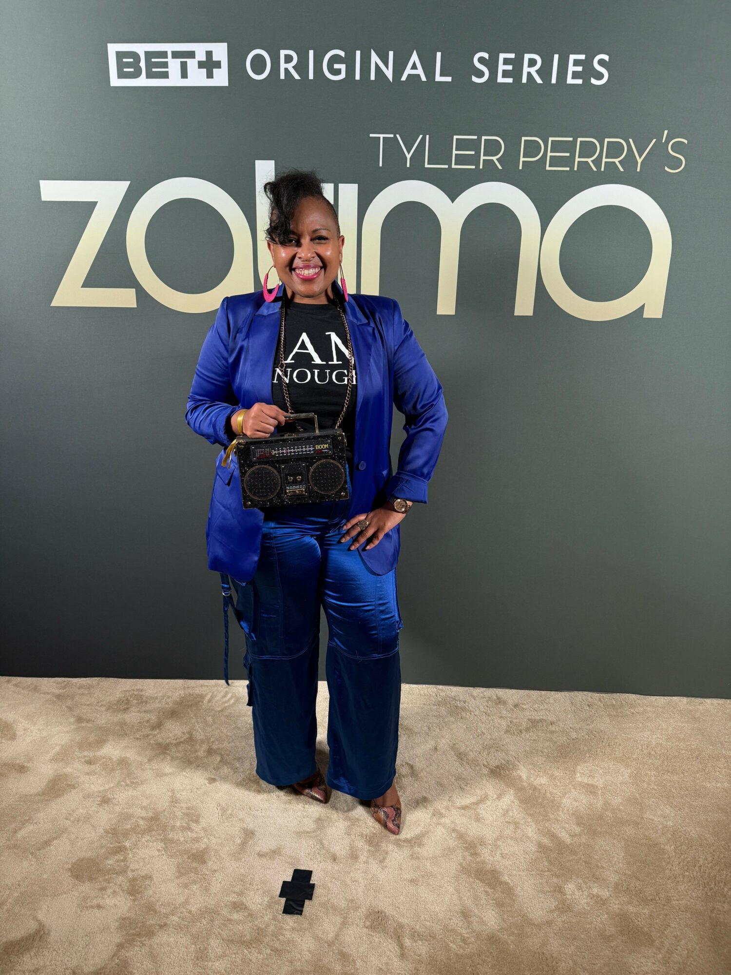 Woman in blue suit holding a camera, standing in front of a promotional backdrop for Tyler Perry's Zoom, smiling.