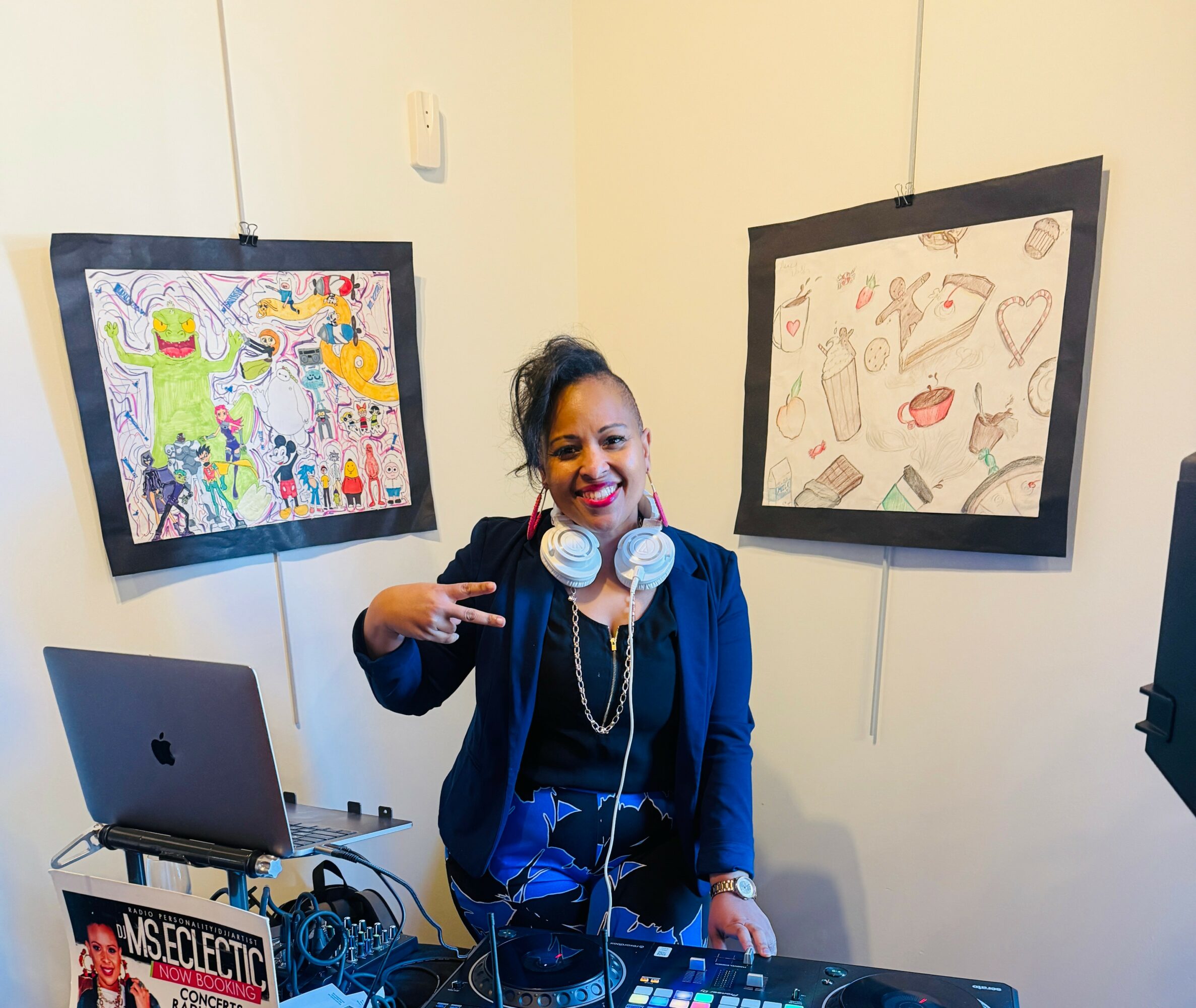 Woman with headphones smiling, pointing at artwork on wall, in a room with a laptop and art supplies.