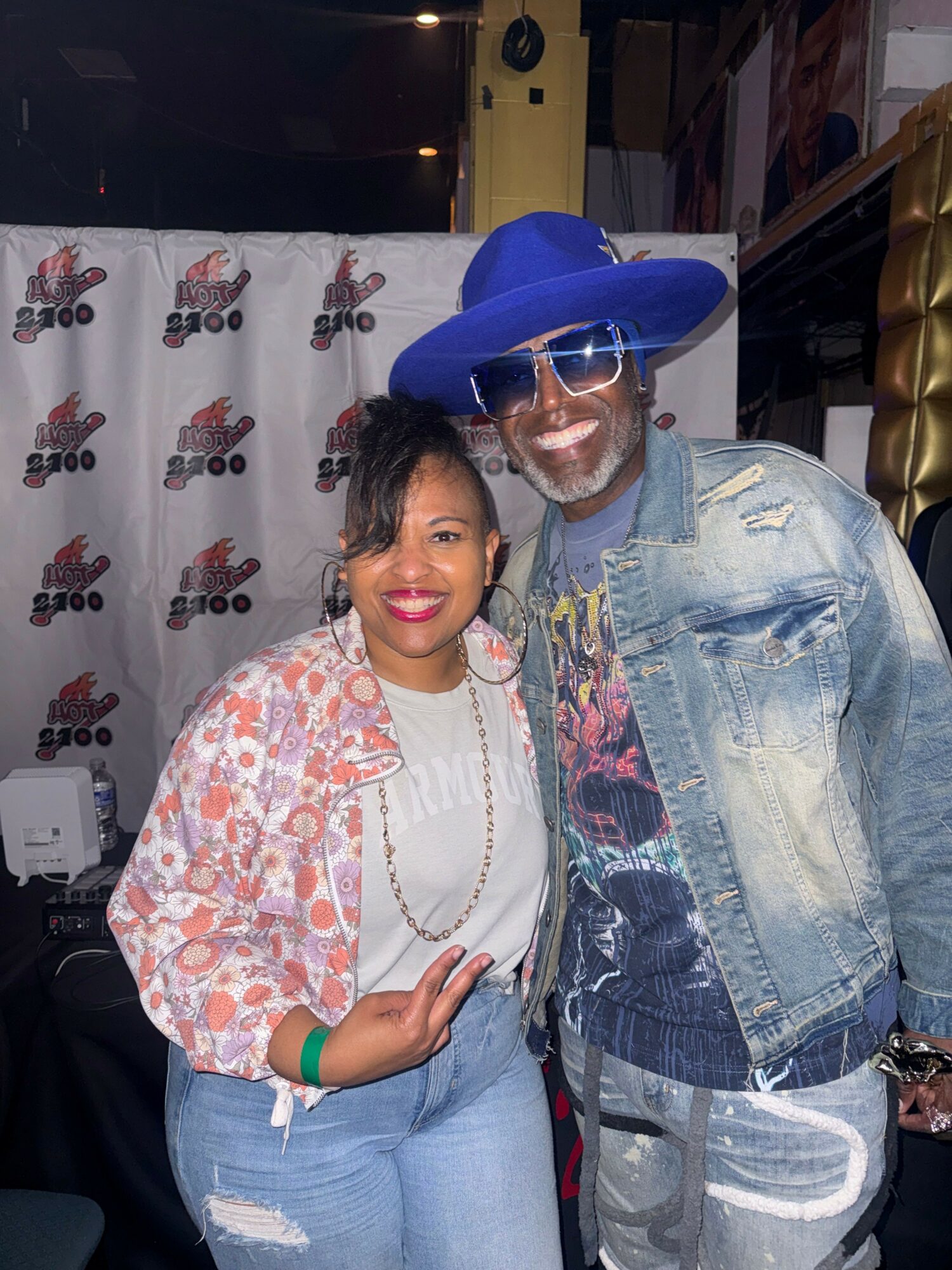 Two people smiling, one wearing a large blue hat and sunglasses, the other in a patterned jacket, in front of a backdrop.
