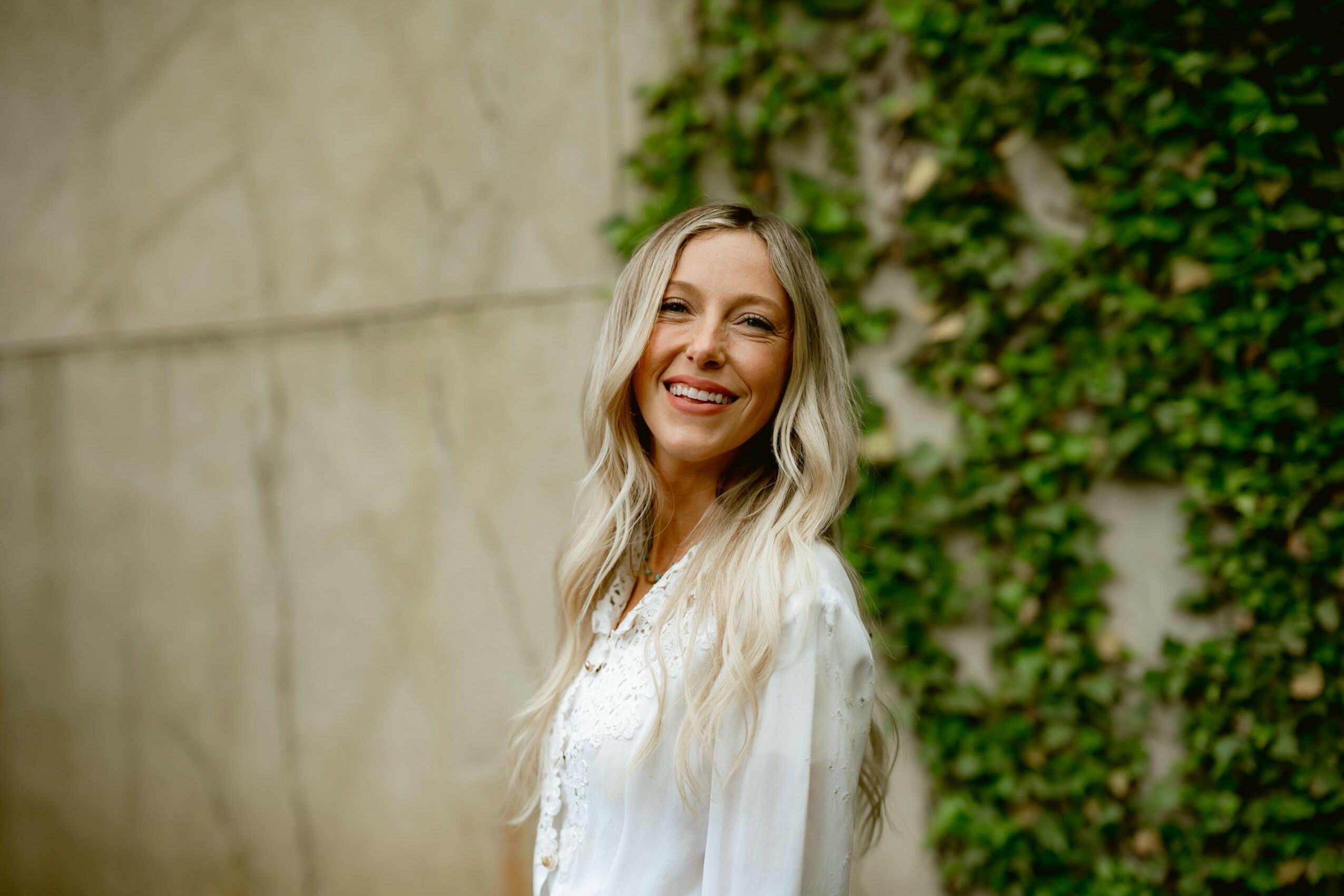 Woman with long wavy blonde hair smiling outdoors near a wall and green ivy.