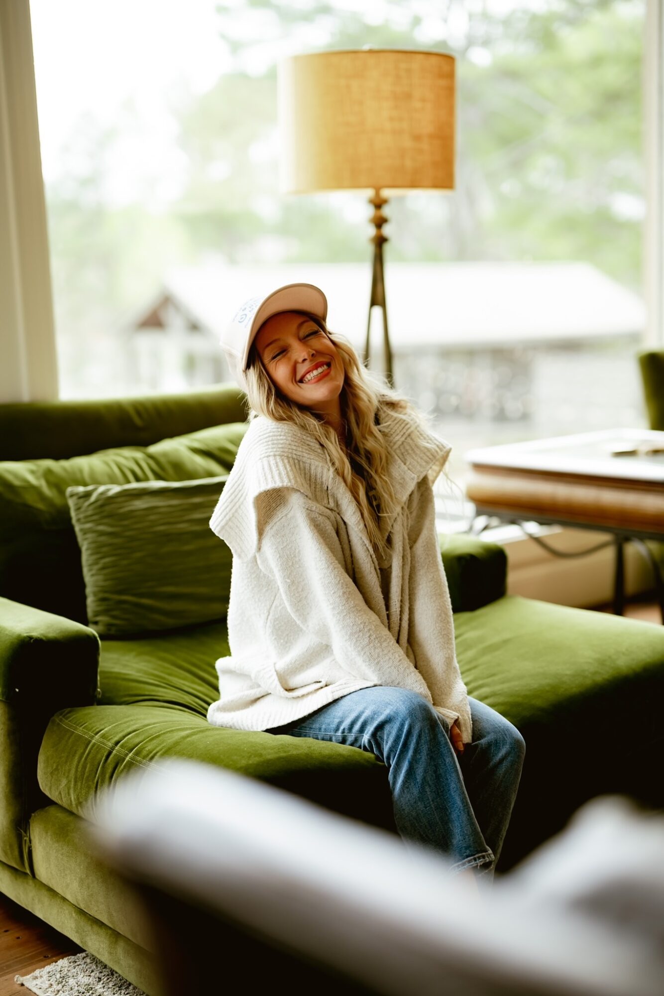 Woman sitting on a green sofa, smiling, wearing a beige hat and oversized sweater, in a bright room with a lamp and window.