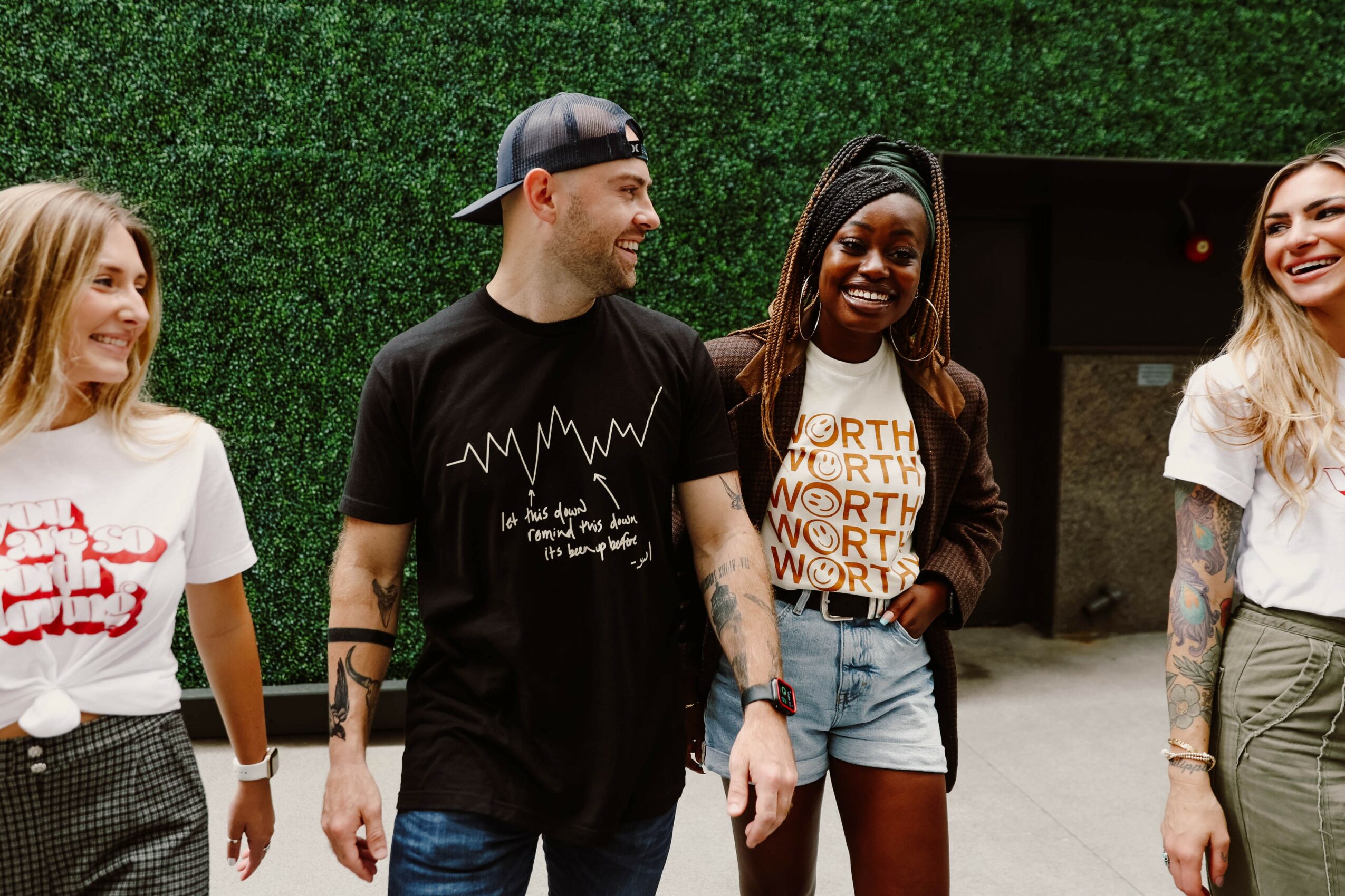 Four people standing outdoors, smiling and talking, with green hedge background.