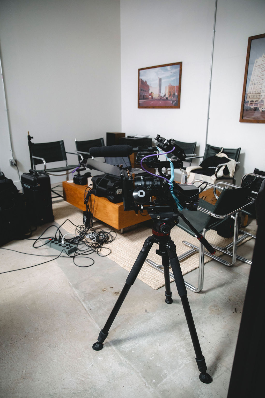 Room with chairs, a camera on a tripod, and electronic equipment, with framed pictures on the wall.