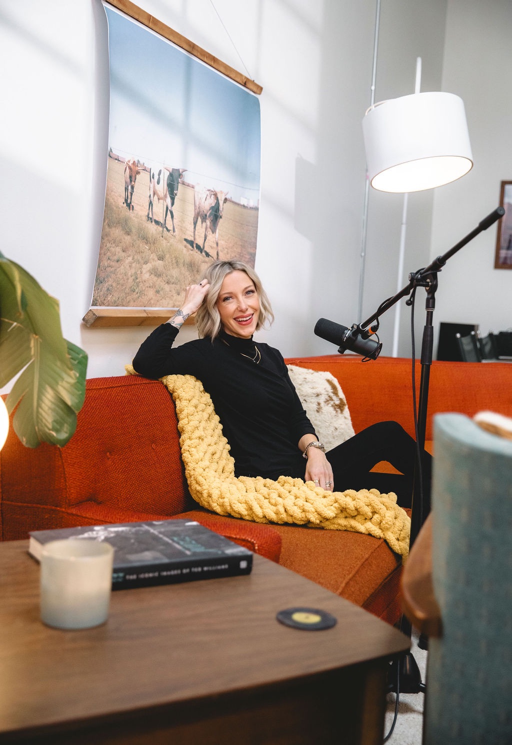 Woman sitting on orange sofa with yellow blanket, smiling, in a room with a large painting of horses, microphone nearby.