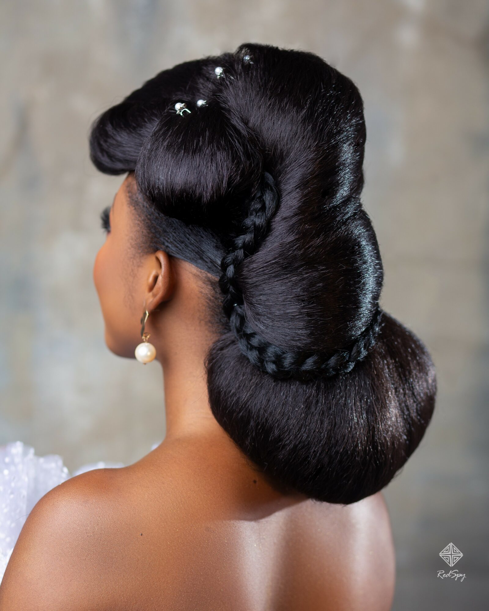 Woman with elaborate vintage hairstyle featuring large, smooth rolls and a braided section, adorned with small decorative flowers.