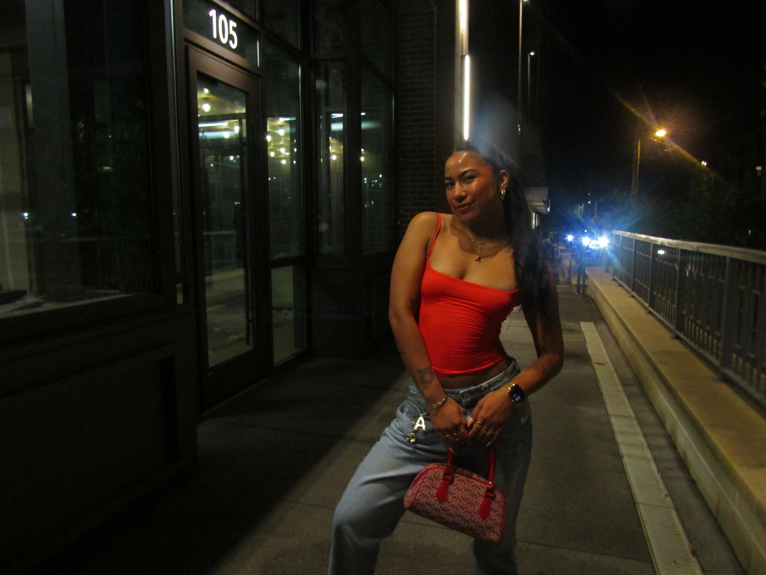 Woman in red top and jeans holding a pink purse standing outside at night near a building and railing.