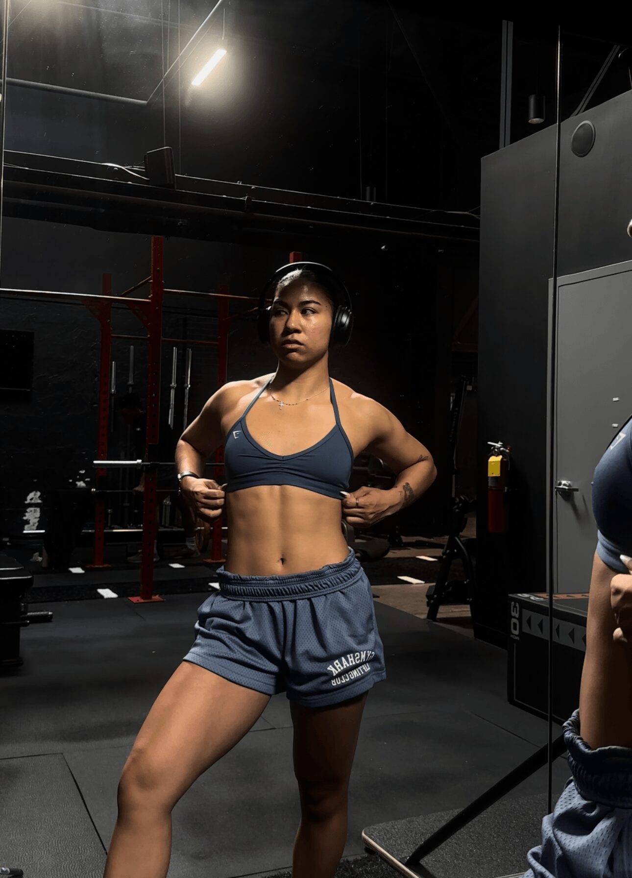 Young woman in workout clothes with headphones in gym, standing with hands on hips.