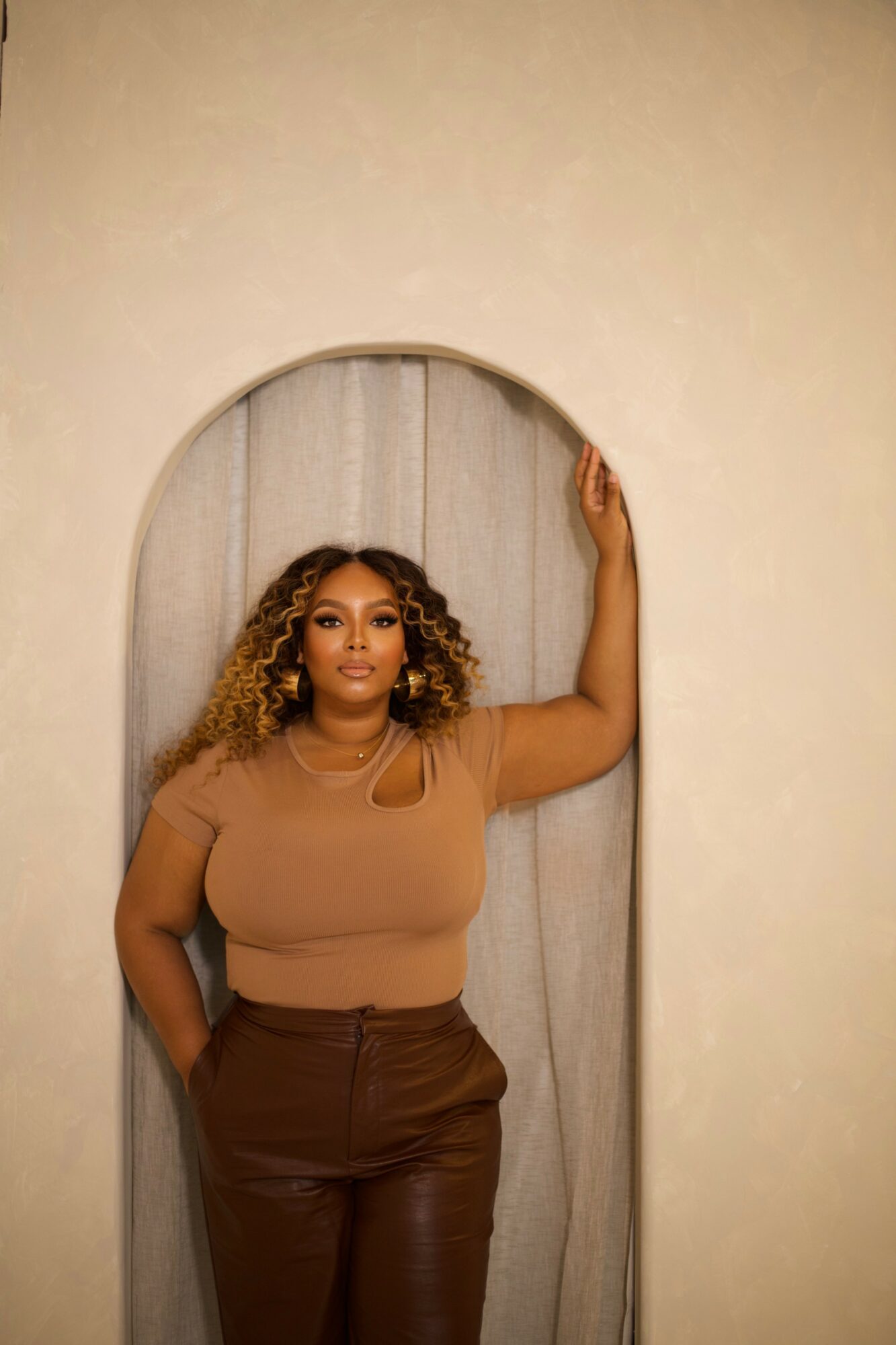 Woman with curly hair in a brown top and pants, standing in an arched doorway, arm raised, neutral expression.