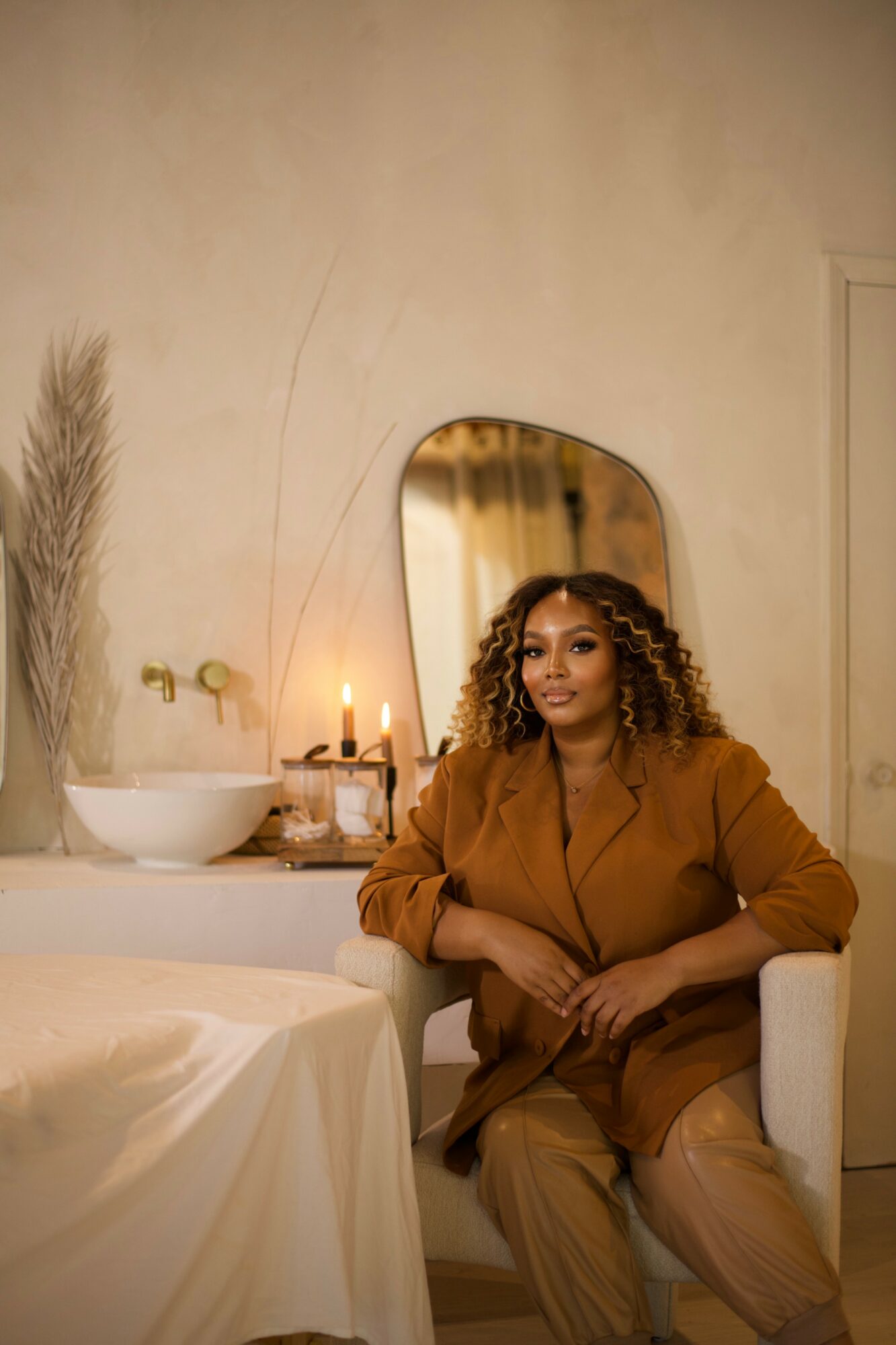 Woman with curly hair sitting on a chair in a room with a mirror, candles, and decorative items on a table.