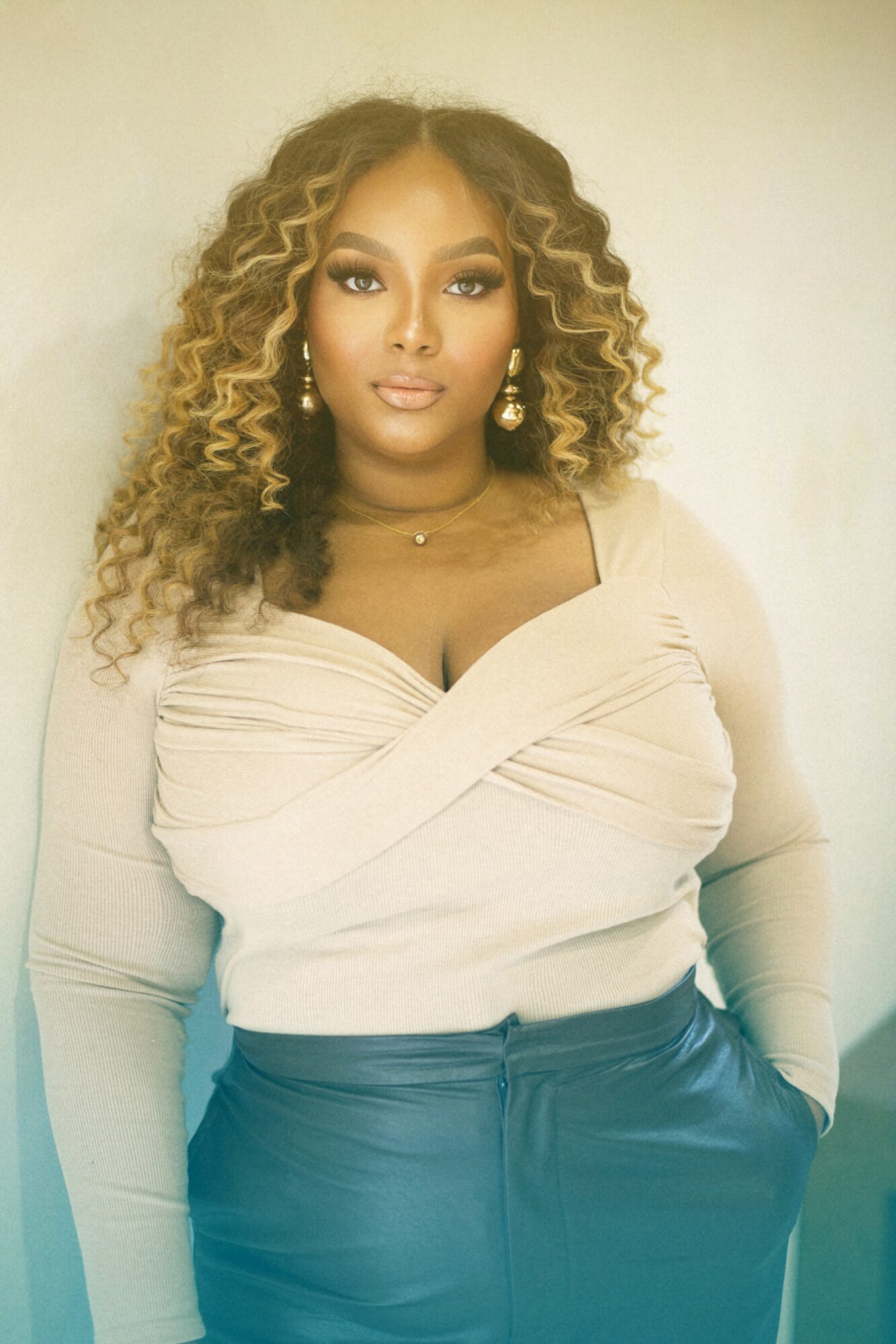 Woman with curly hair, wearing a beige top and dark pants, posing indoors against a plain background.