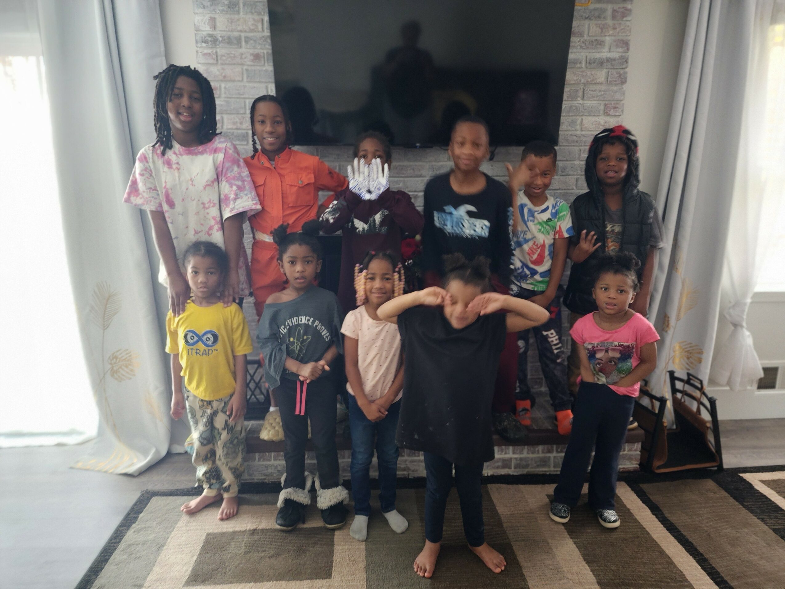 Group of twelve children standing indoors in front of a TV, some smiling, some making gestures, diverse in clothing and appearance.