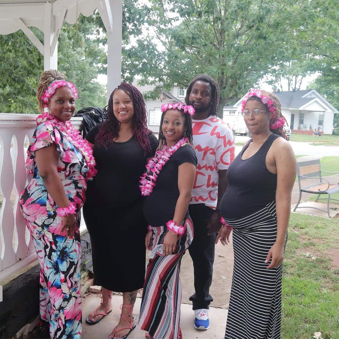Group of five women standing outdoors on a porch with trees and houses in background, smiling at camera.