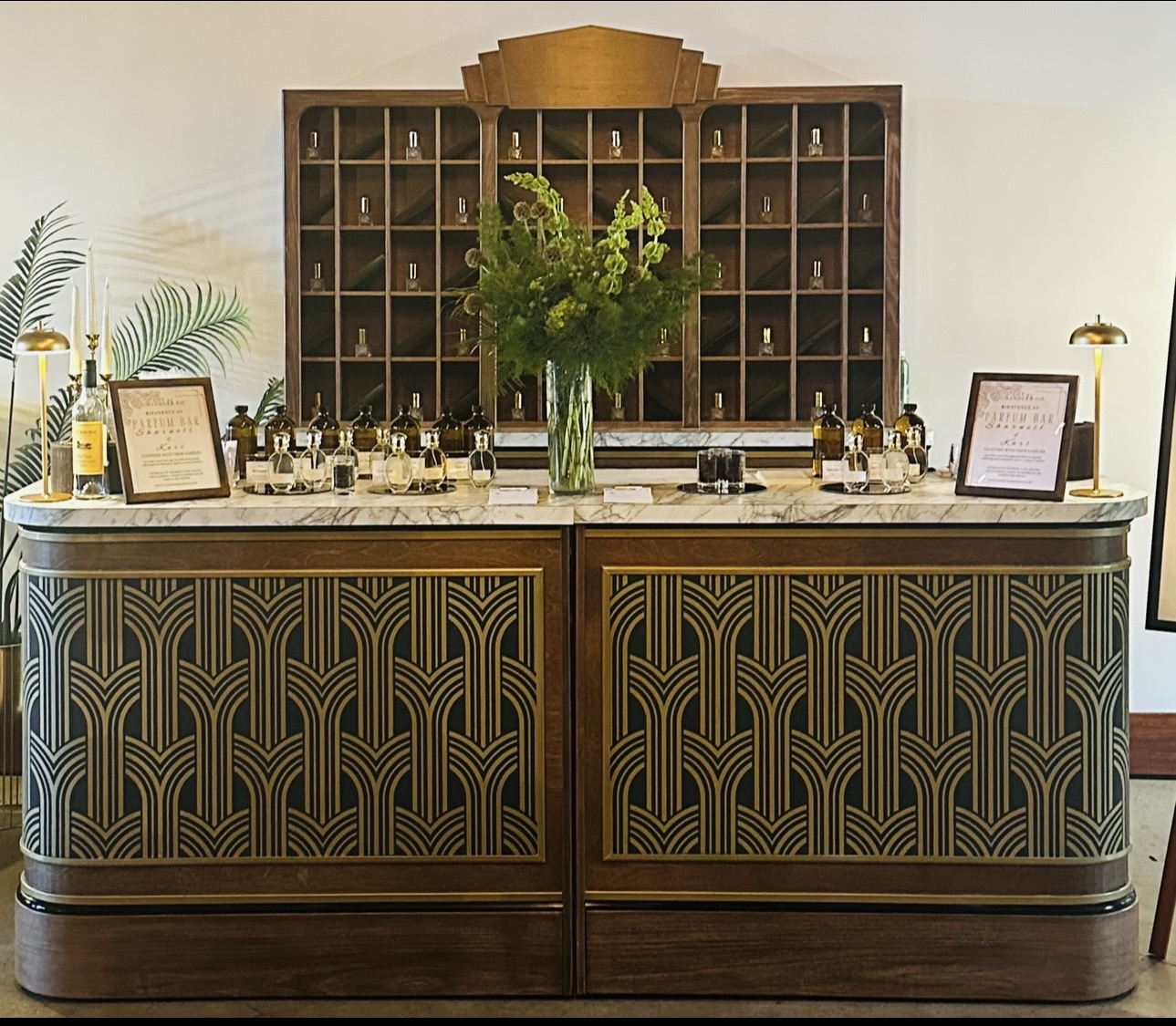 Counter with a large vase of green flowers, framed signs, and small bottles, with a wooden shelf behind displaying bottles.