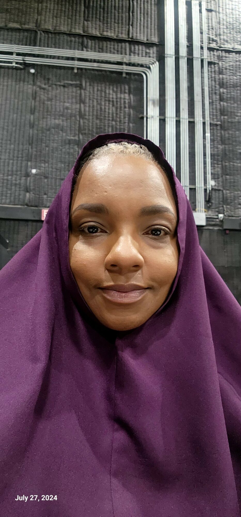 Woman wearing a purple headscarf, smiling, in an industrial setting with metal pipes and walls.