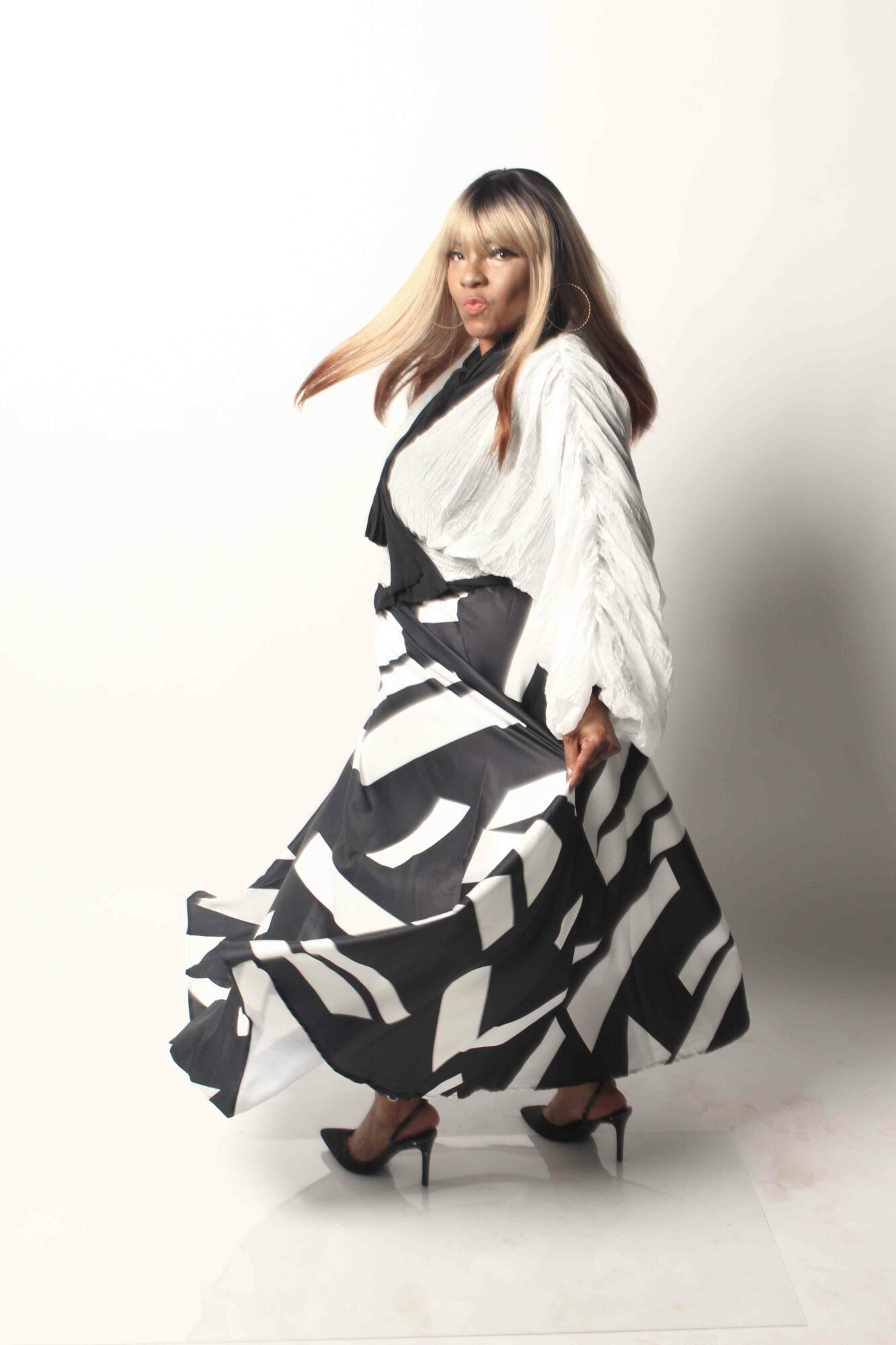 Woman in black and white patterned dress and white jacket, standing against a plain white background.