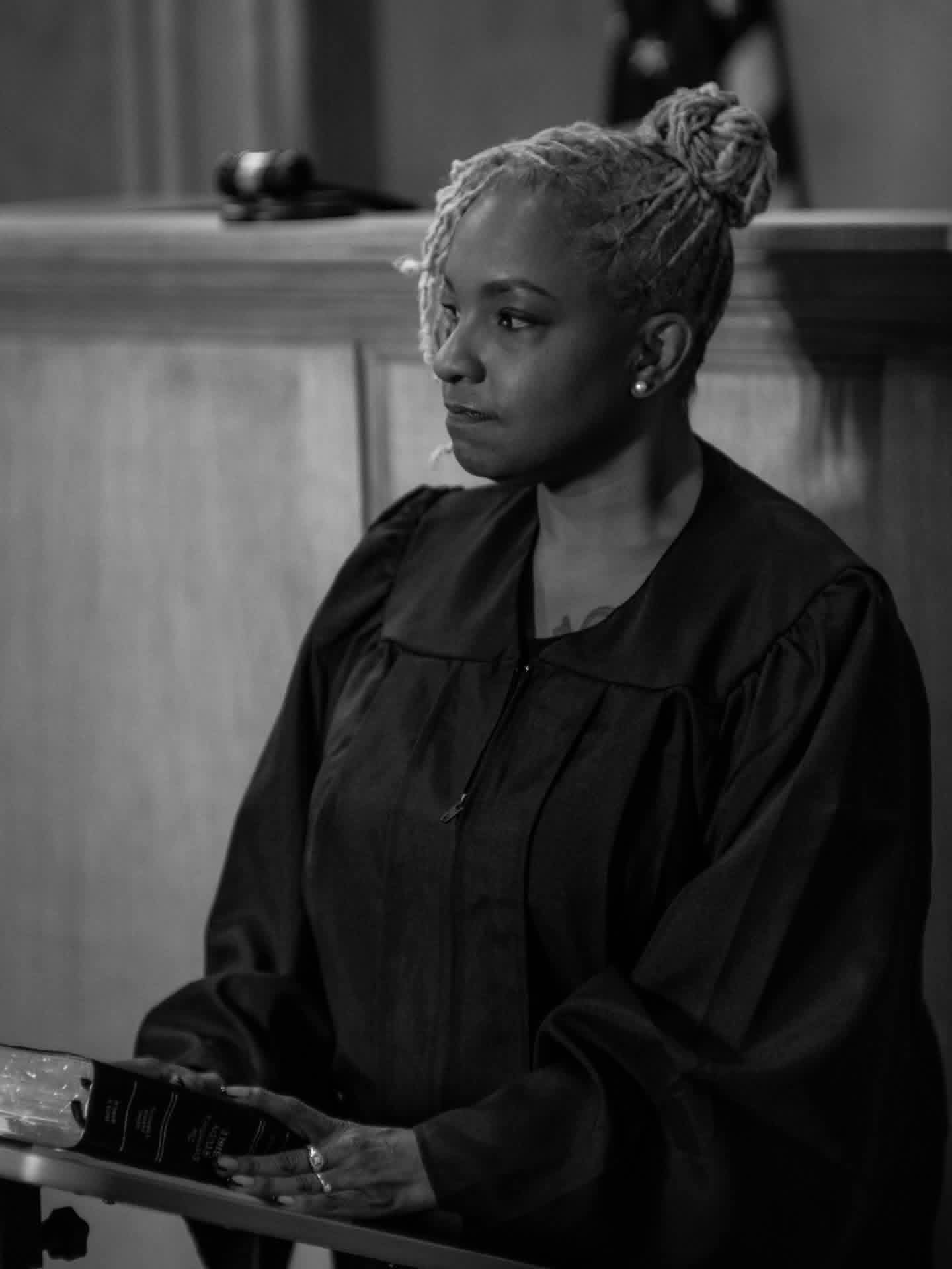 Woman with braided hair and earrings, wearing a black robe, sitting in a courtroom or formal setting.