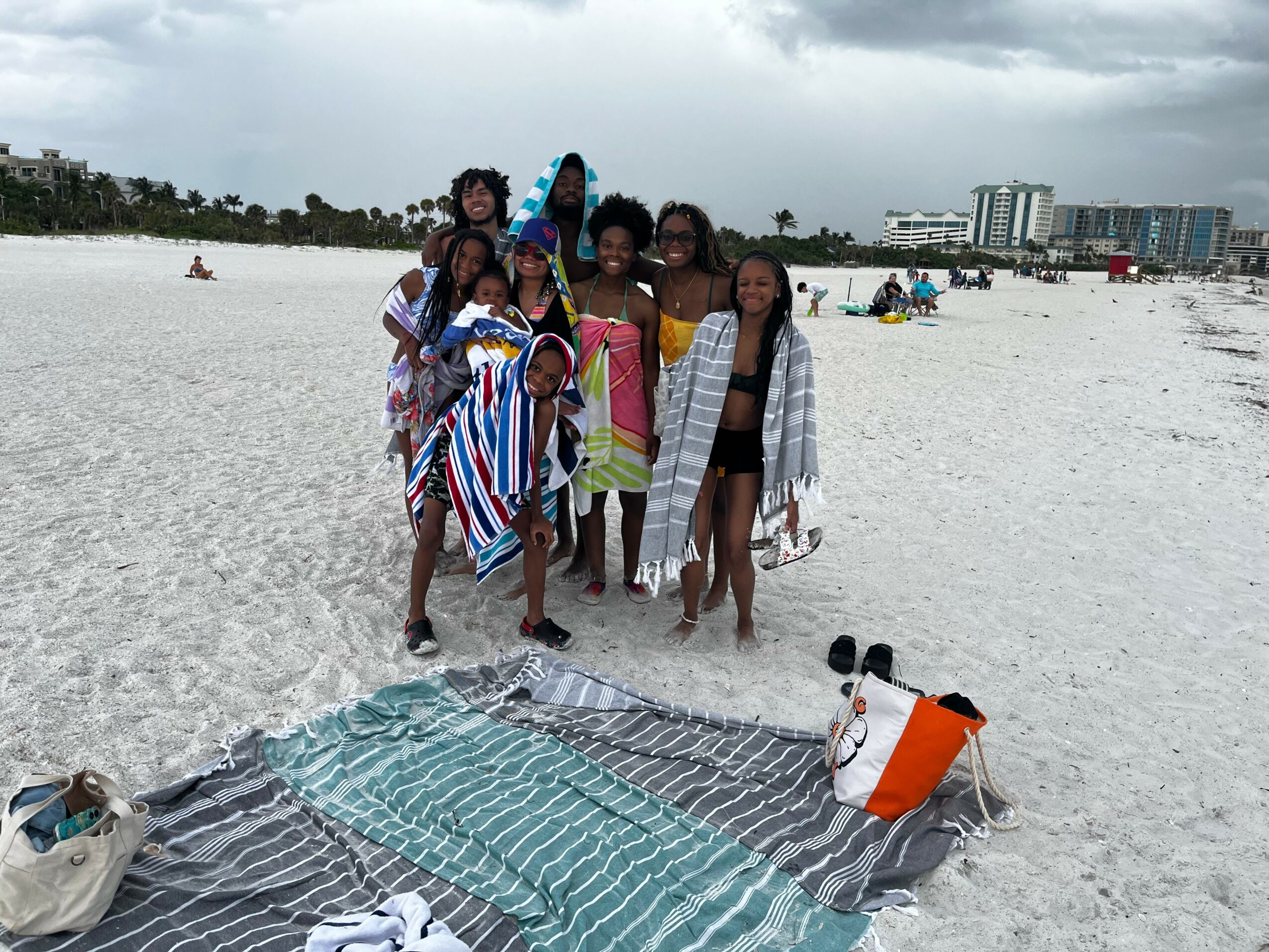 Group of children standing on a sandy beach with a cityscape in the background, some holding umbrellas and towels.