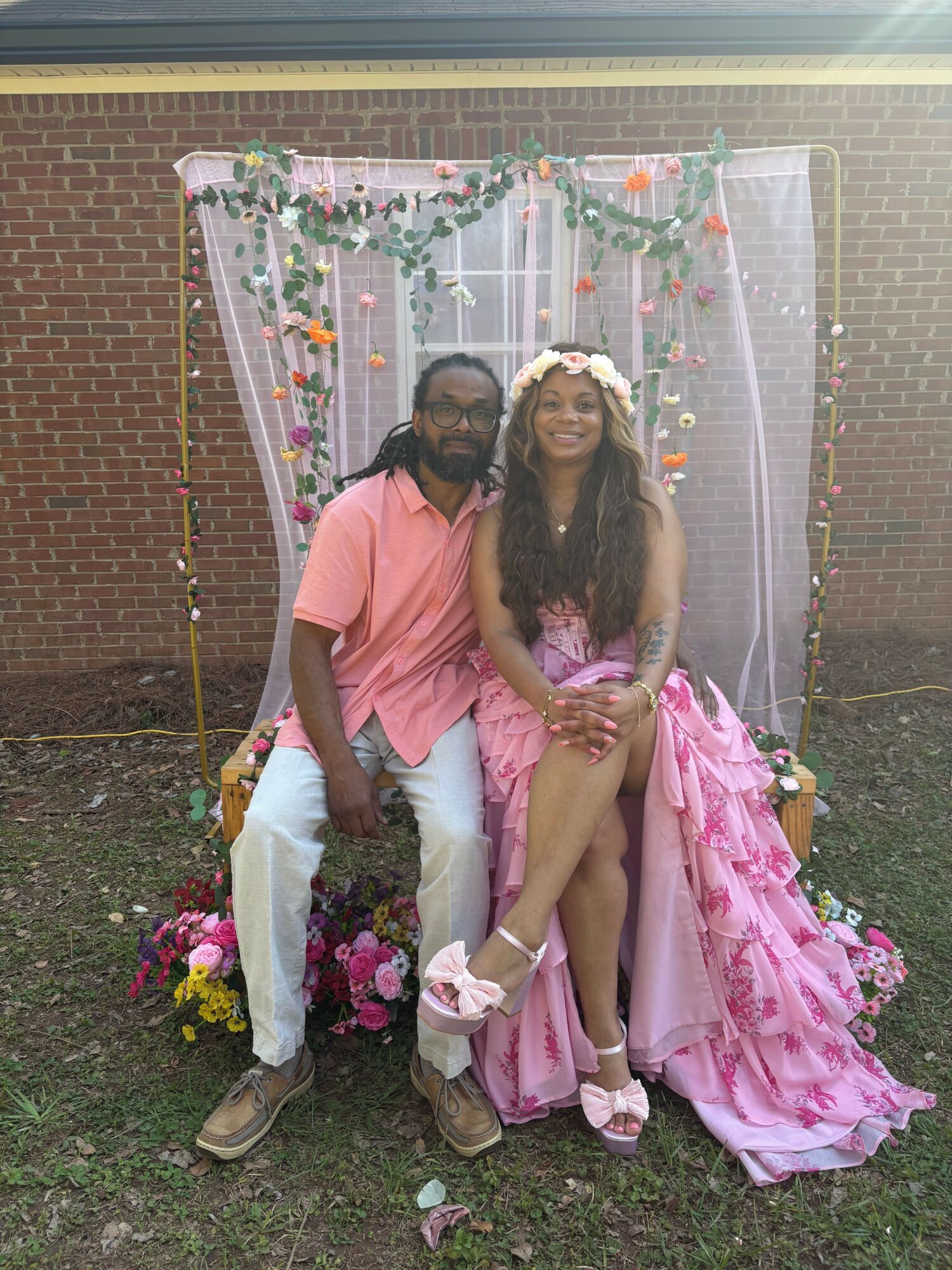 Two people sitting on a decorated chair in front of a floral backdrop, smiling, outdoors, brick wall background.