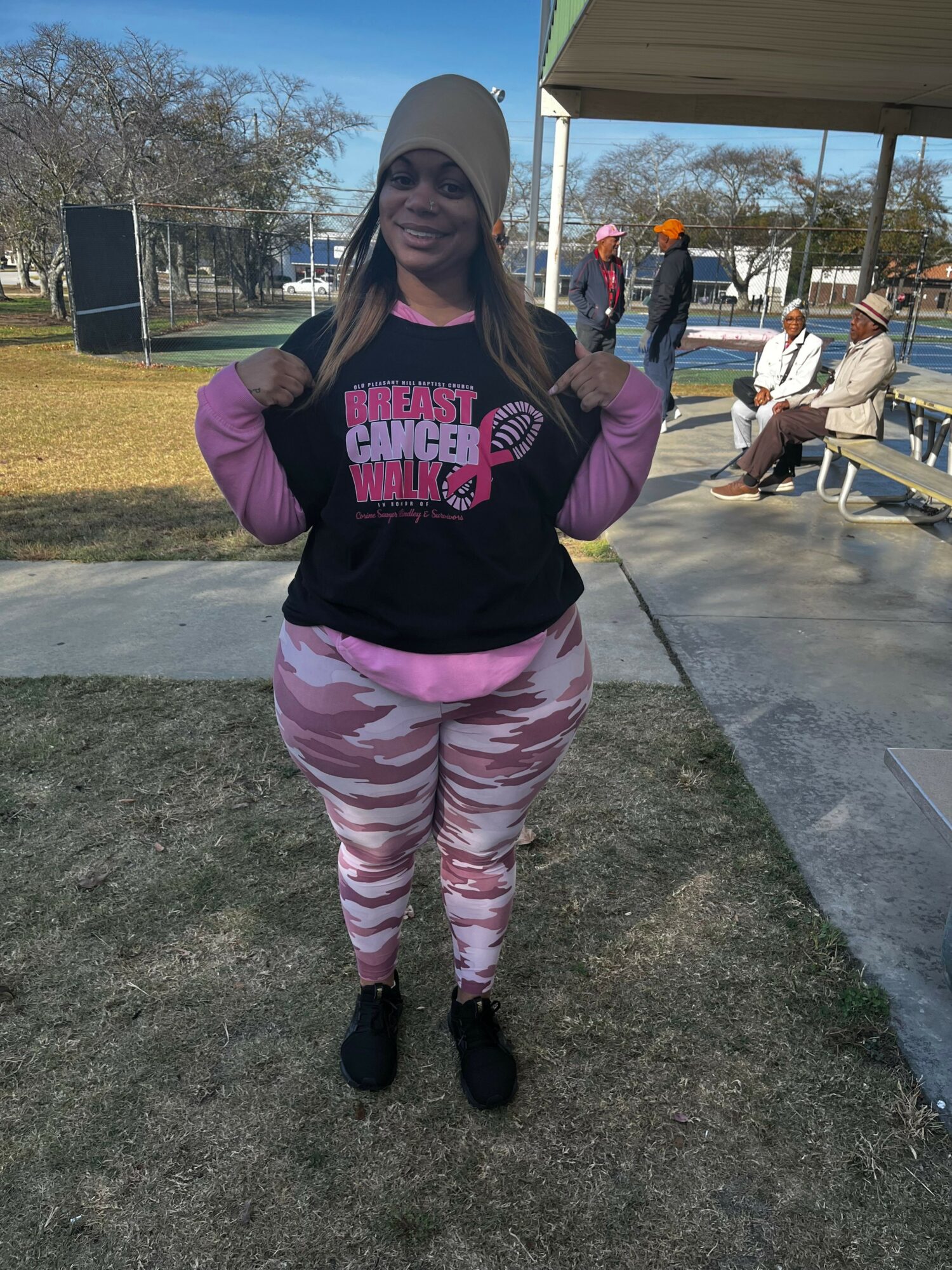 Young woman standing outdoors, smiling, wearing a beanie, black hoodie, pink camouflage pants, and black shoes.