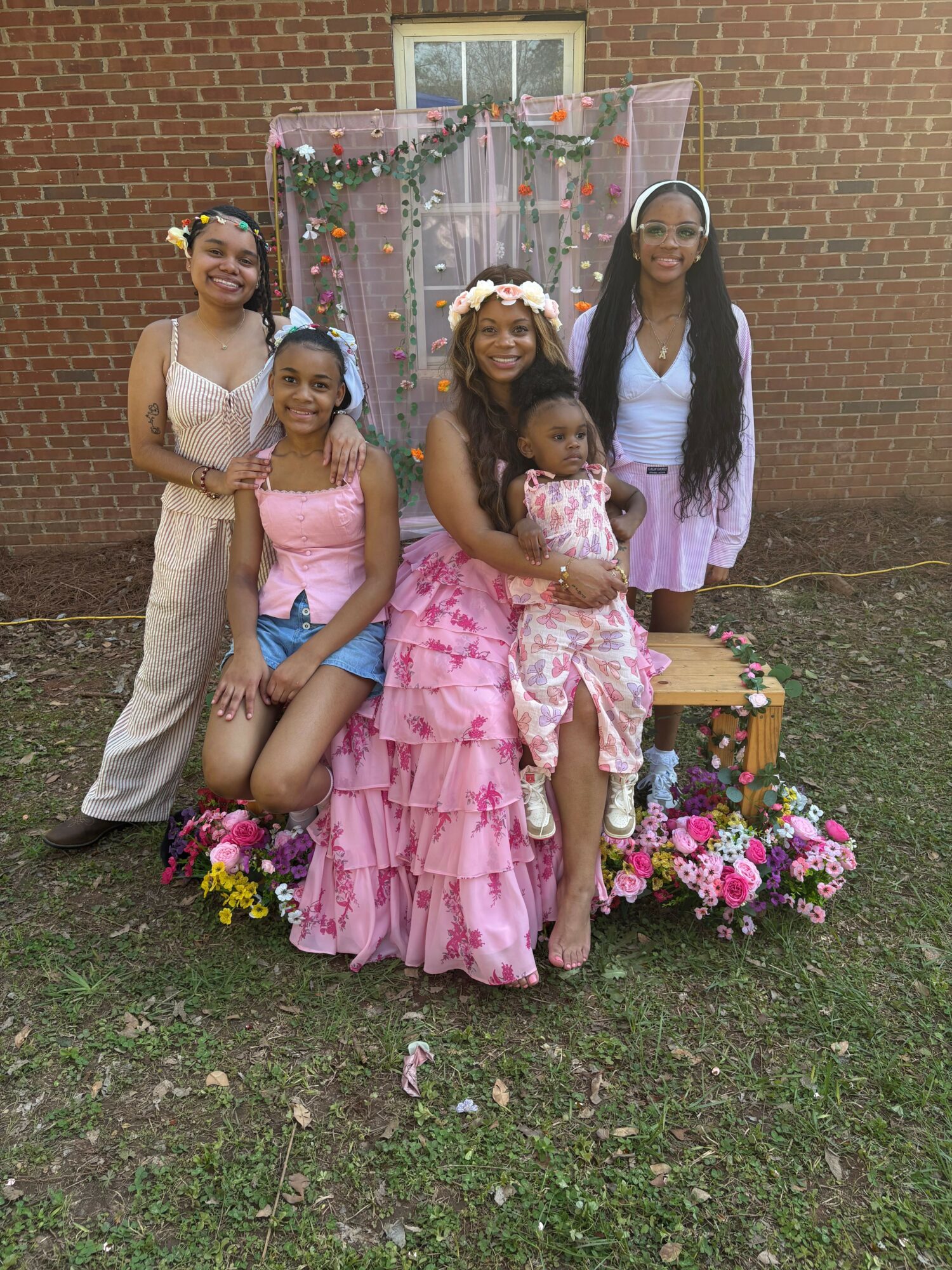 Five girls standing outdoors in front of a decorated backdrop with flowers, smiling, with grass and a brick wall behind.