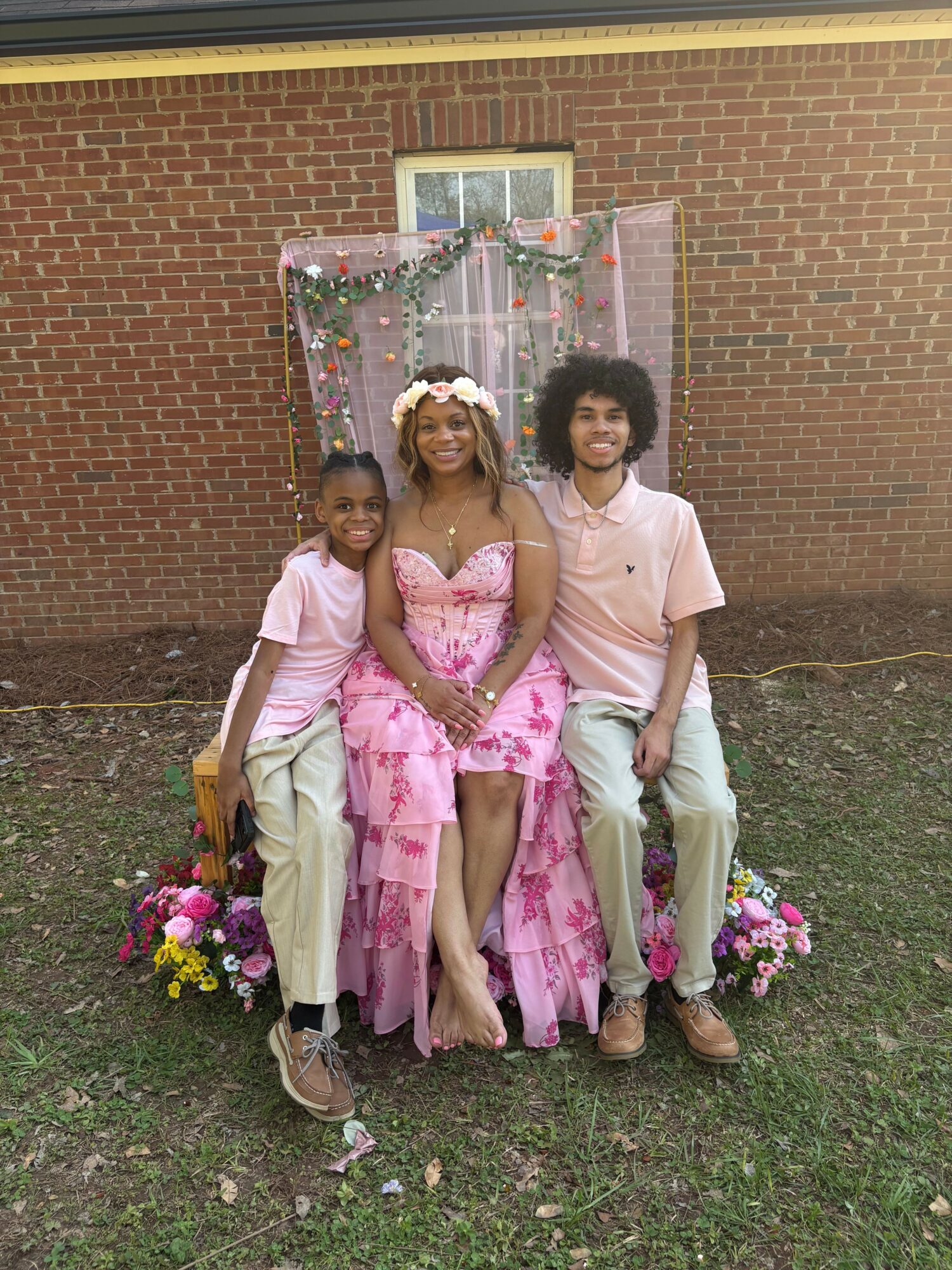 Three people sit on a decorated chair outside a brick building, with flowers on the ground, smiling at the camera.