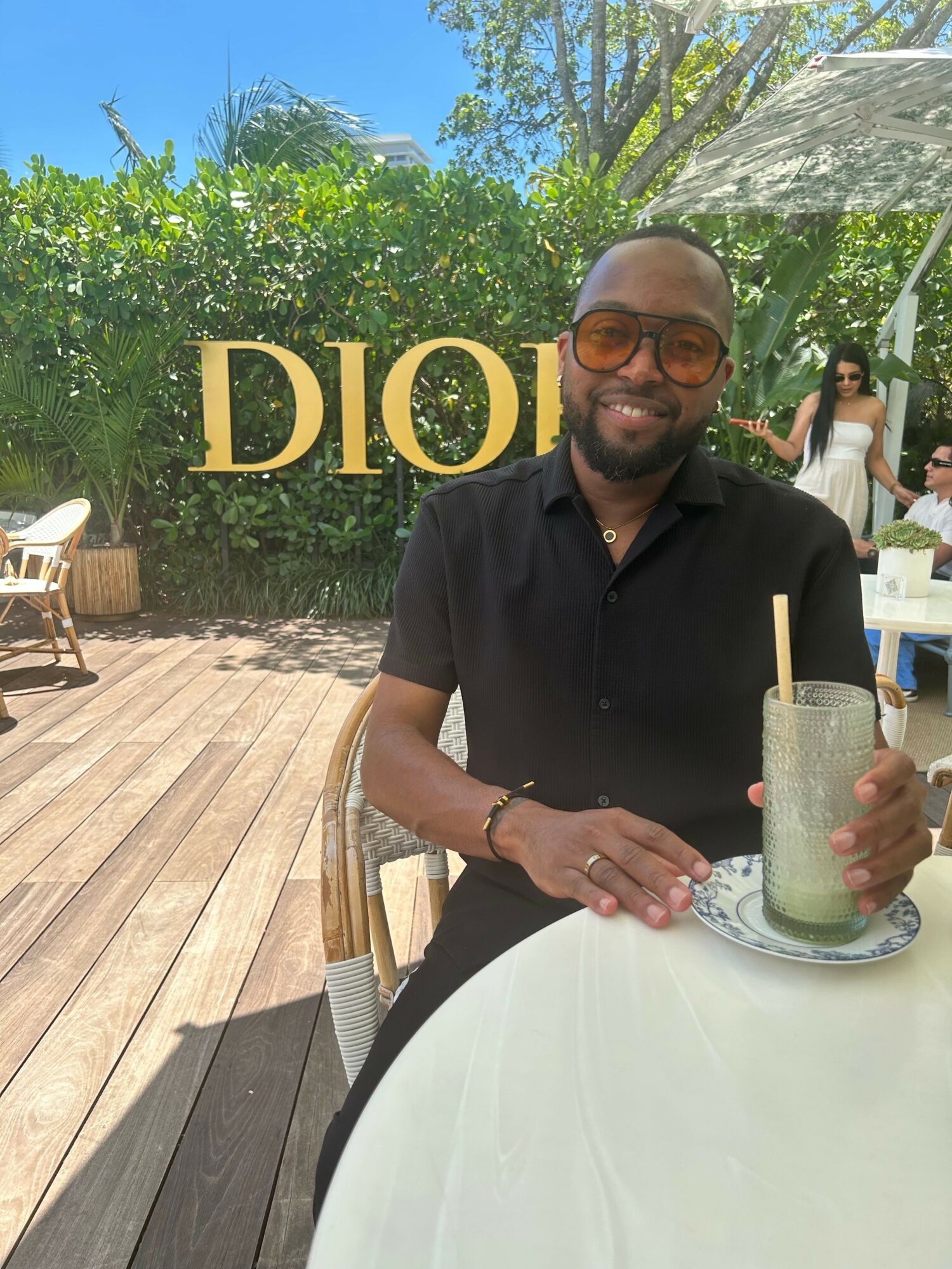 Man with glasses and a beard sitting at a table outdoors, holding a drink with a straw, smiling.