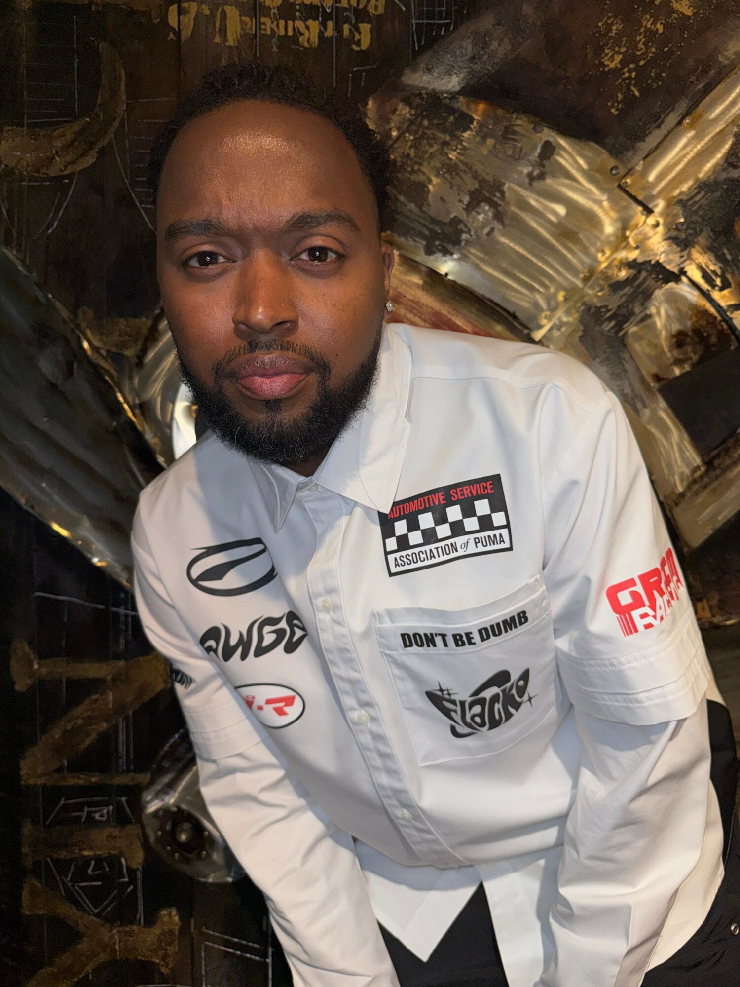 Man with beard and short hair wearing a white racing jacket with logos, standing indoors.