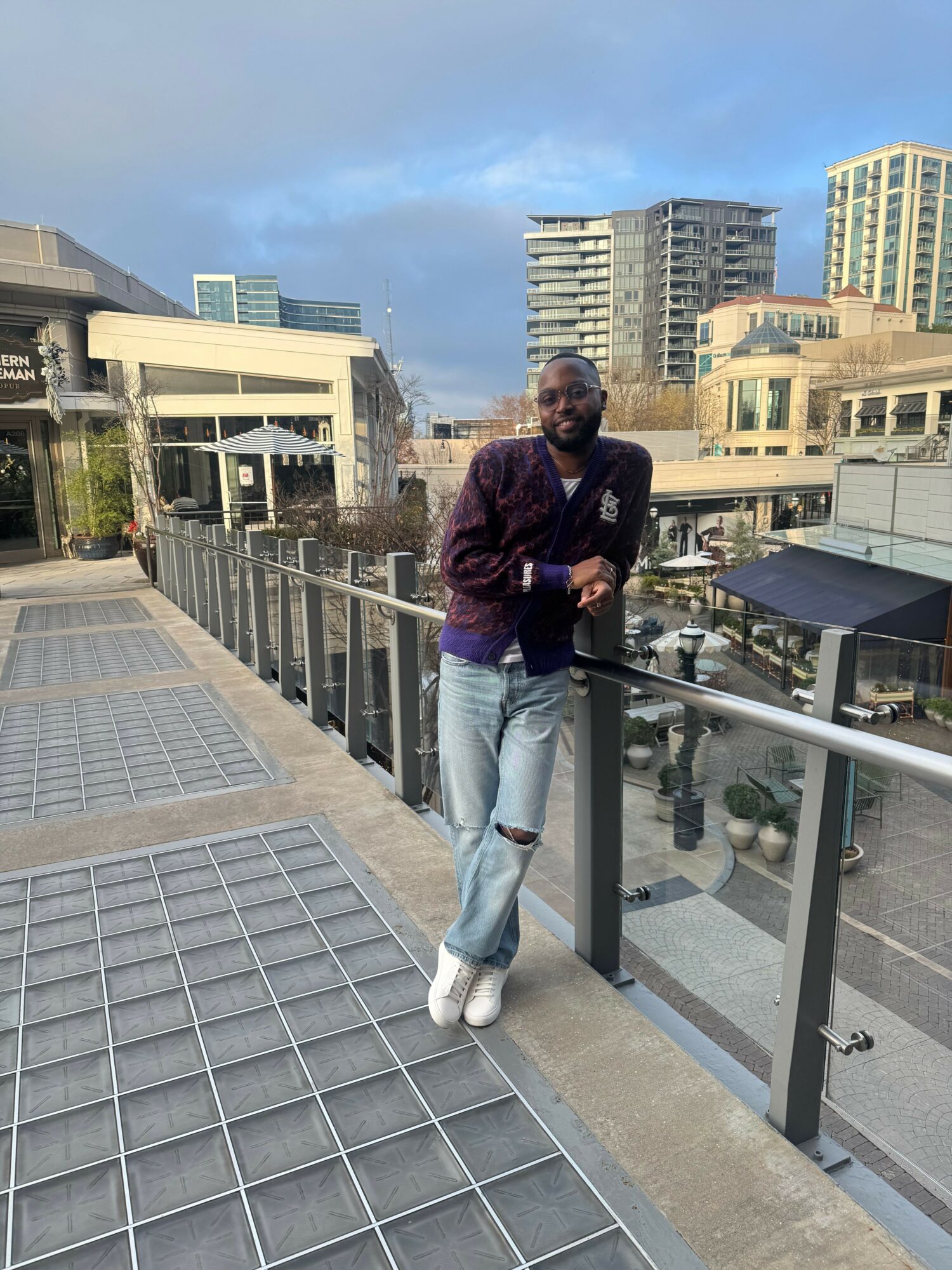 Man standing on a balcony with city buildings in the background, wearing a dark jacket, light jeans, and white shoes.