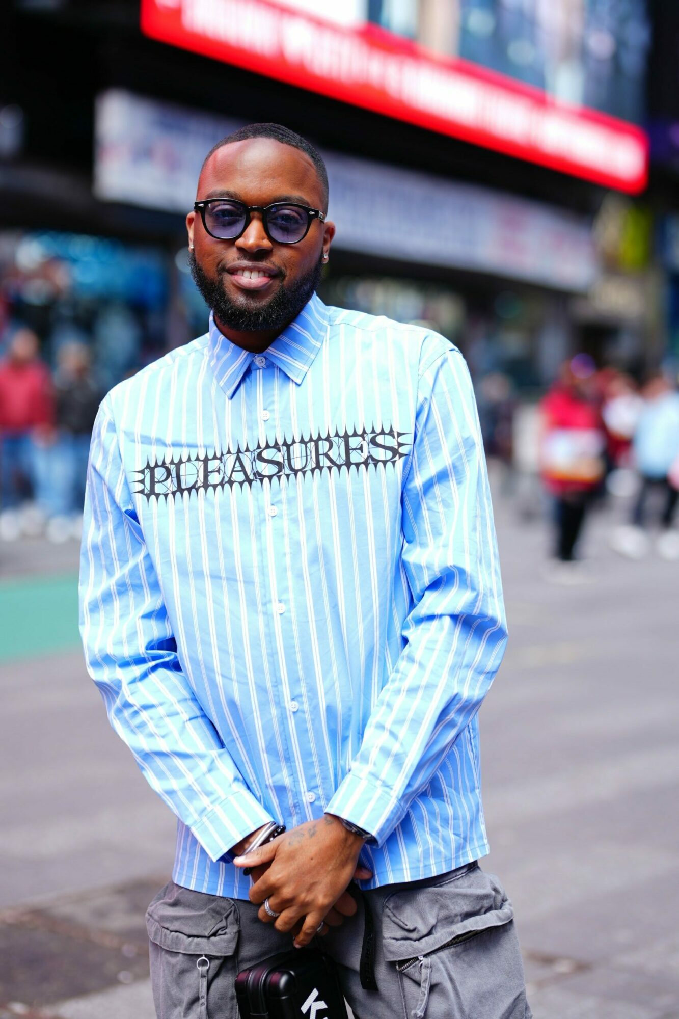 Man wearing sunglasses, blue striped shirt, and gray pants standing outdoors in a busy area.