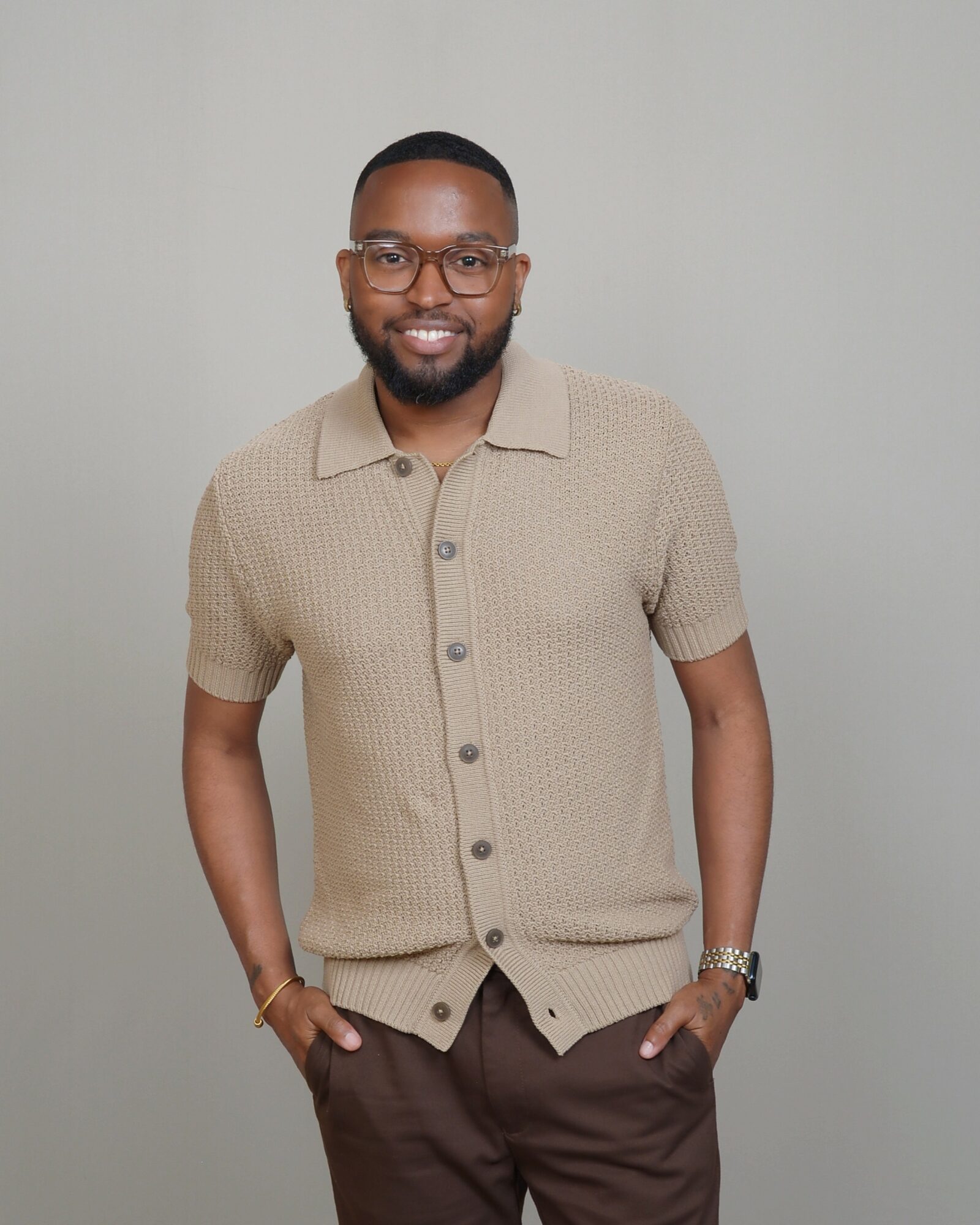 Man with glasses and beard wearing a beige short-sleeve shirt and dark pants, smiling, standing against a plain background.