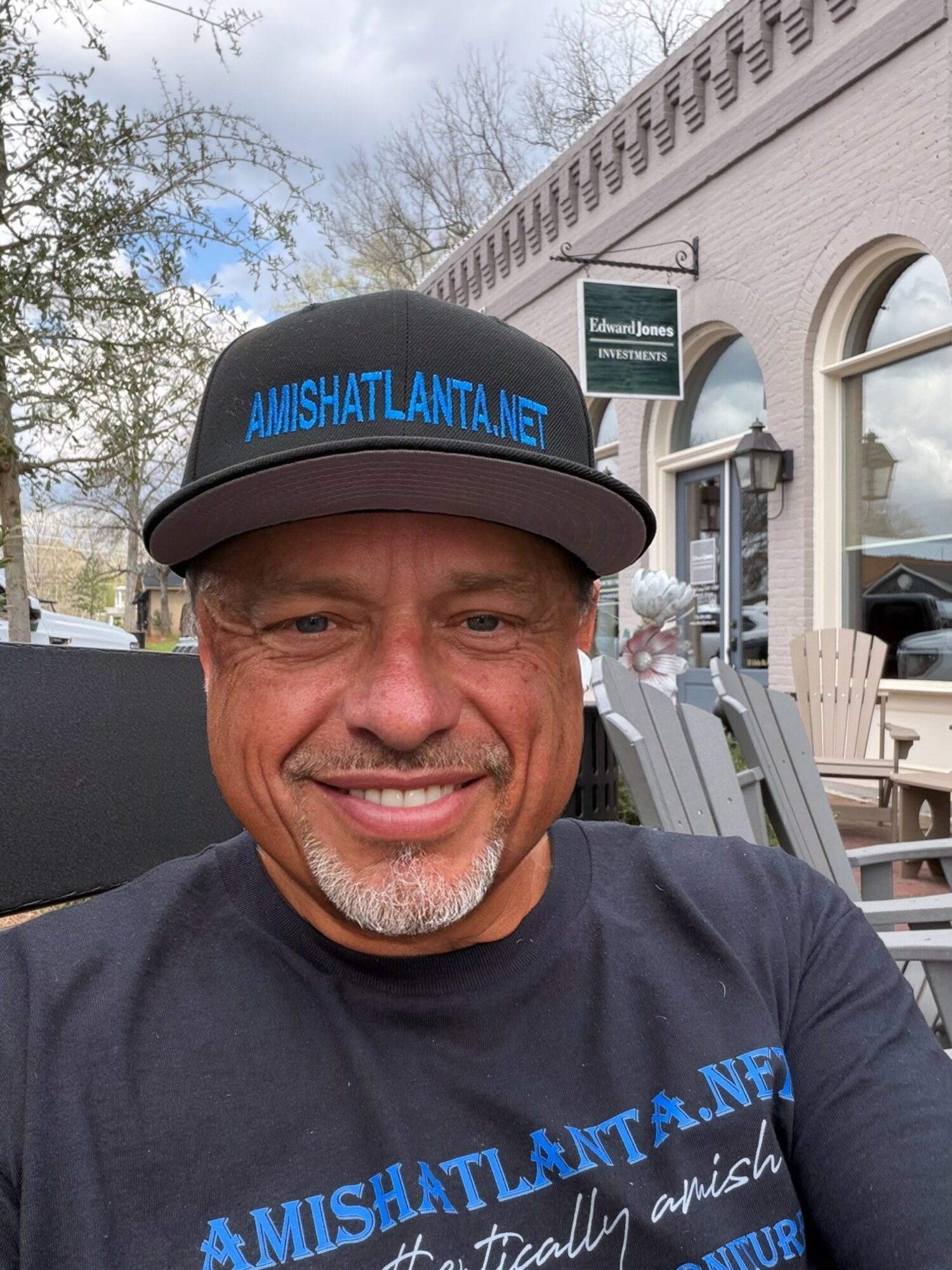 Smiling man wearing a black cap with blue text and a black T-shirt outdoors near a building with large windows.