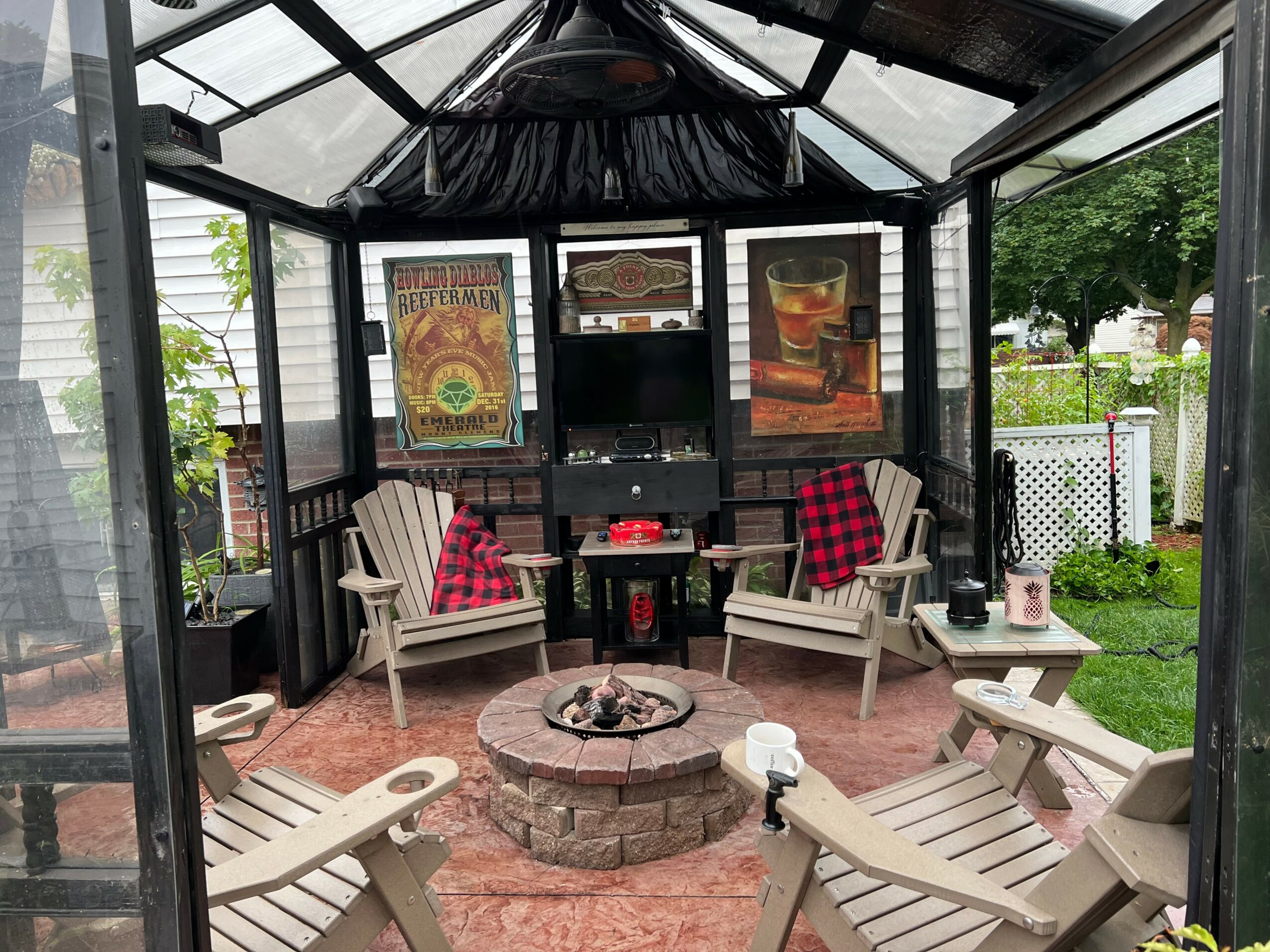 Gazebo with two benches, a fire pit, and two chairs with red cushions, surrounded by a garden and white fence.