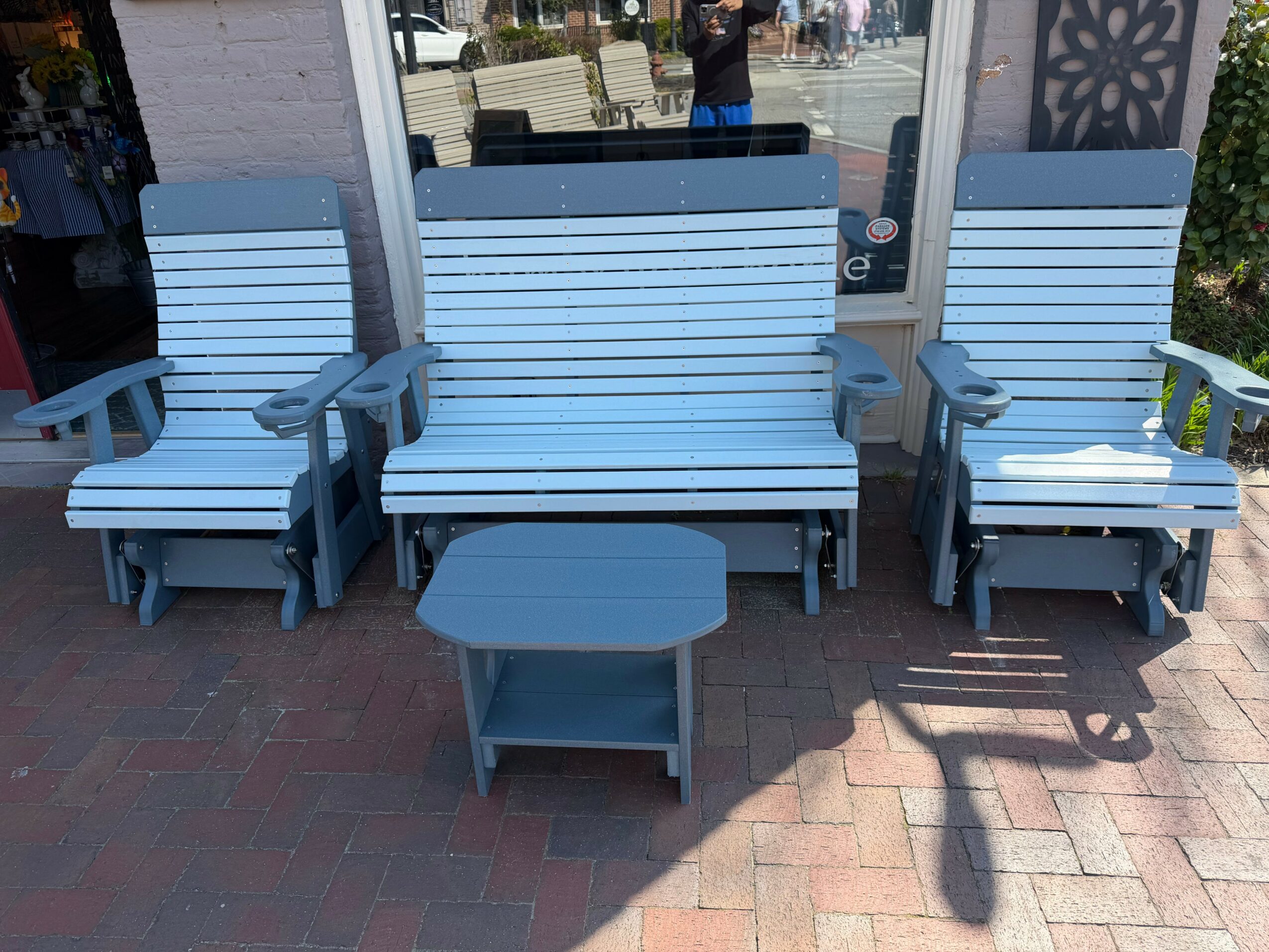 Three white outdoor chairs with armrests and a matching table on a brick sidewalk outside a store.