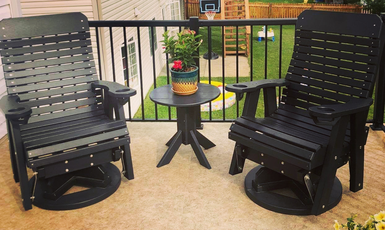 Two black patio chairs with a small round table holding a potted plant on a balcony with a railing and garden in background.