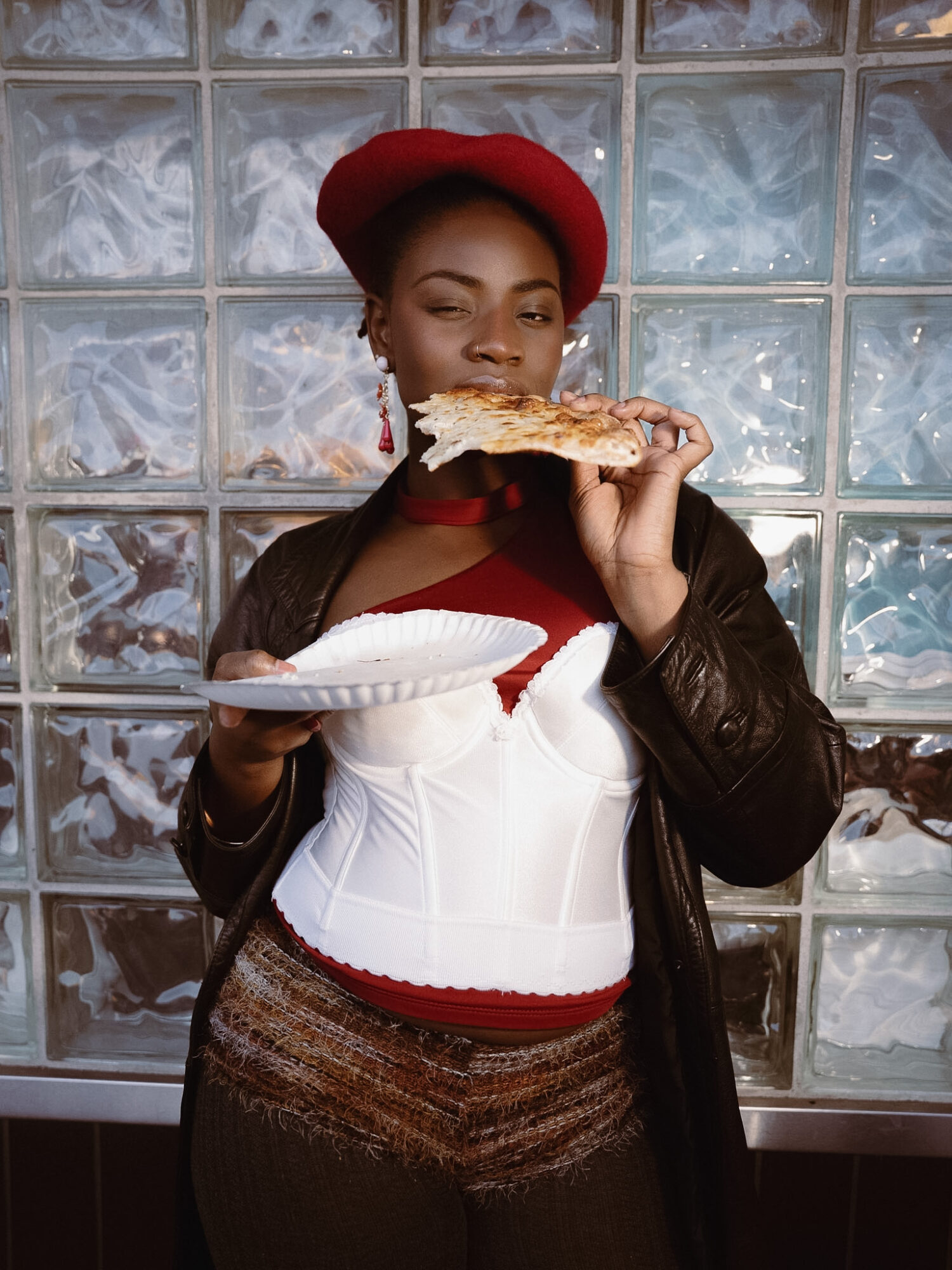 Woman in red hat and leather jacket holding a slice of pizza and a plate, standing in front of glass block wall.